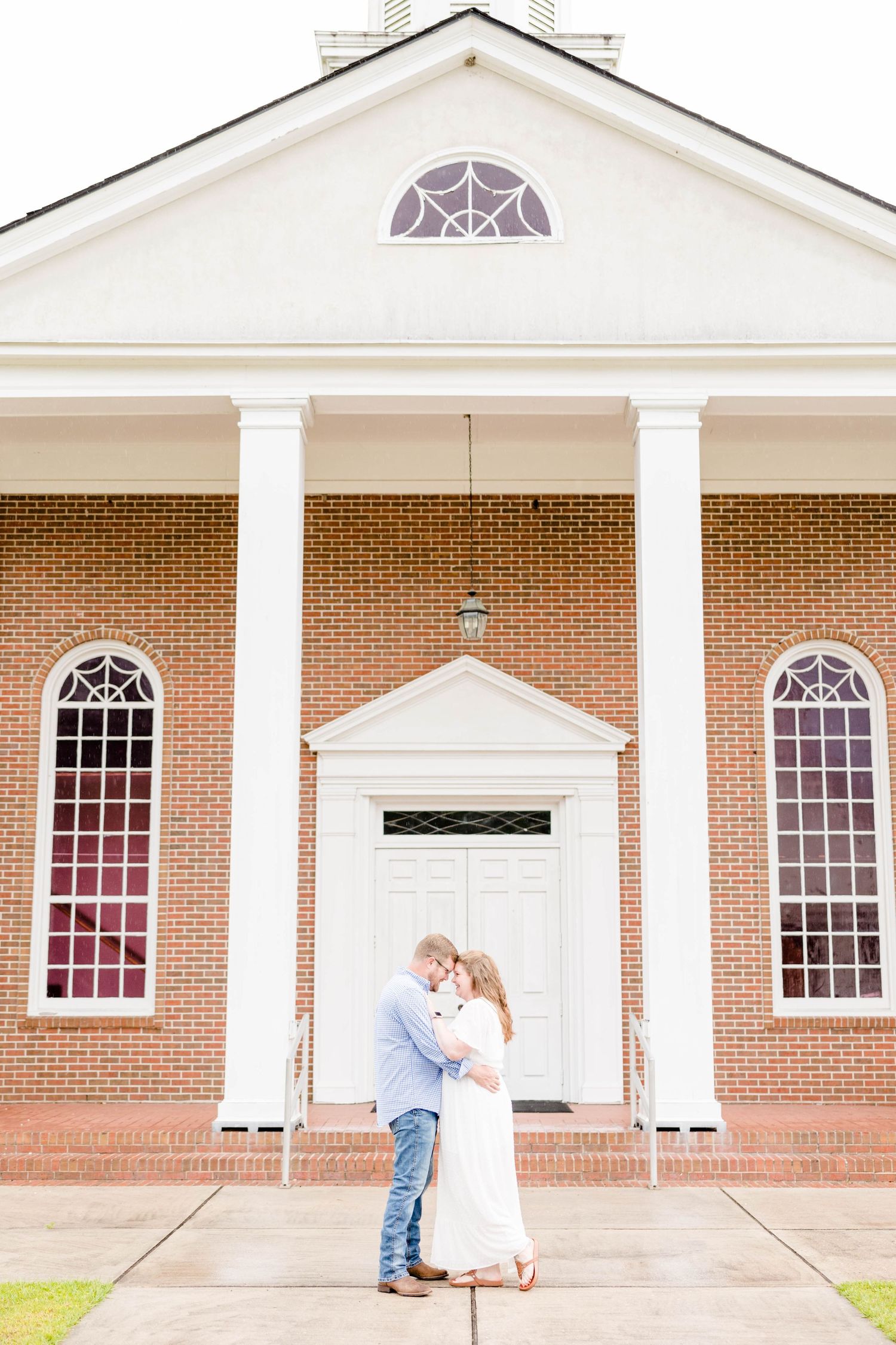 A couple embraces in front of a classic red brick church with white columns and arched windows.