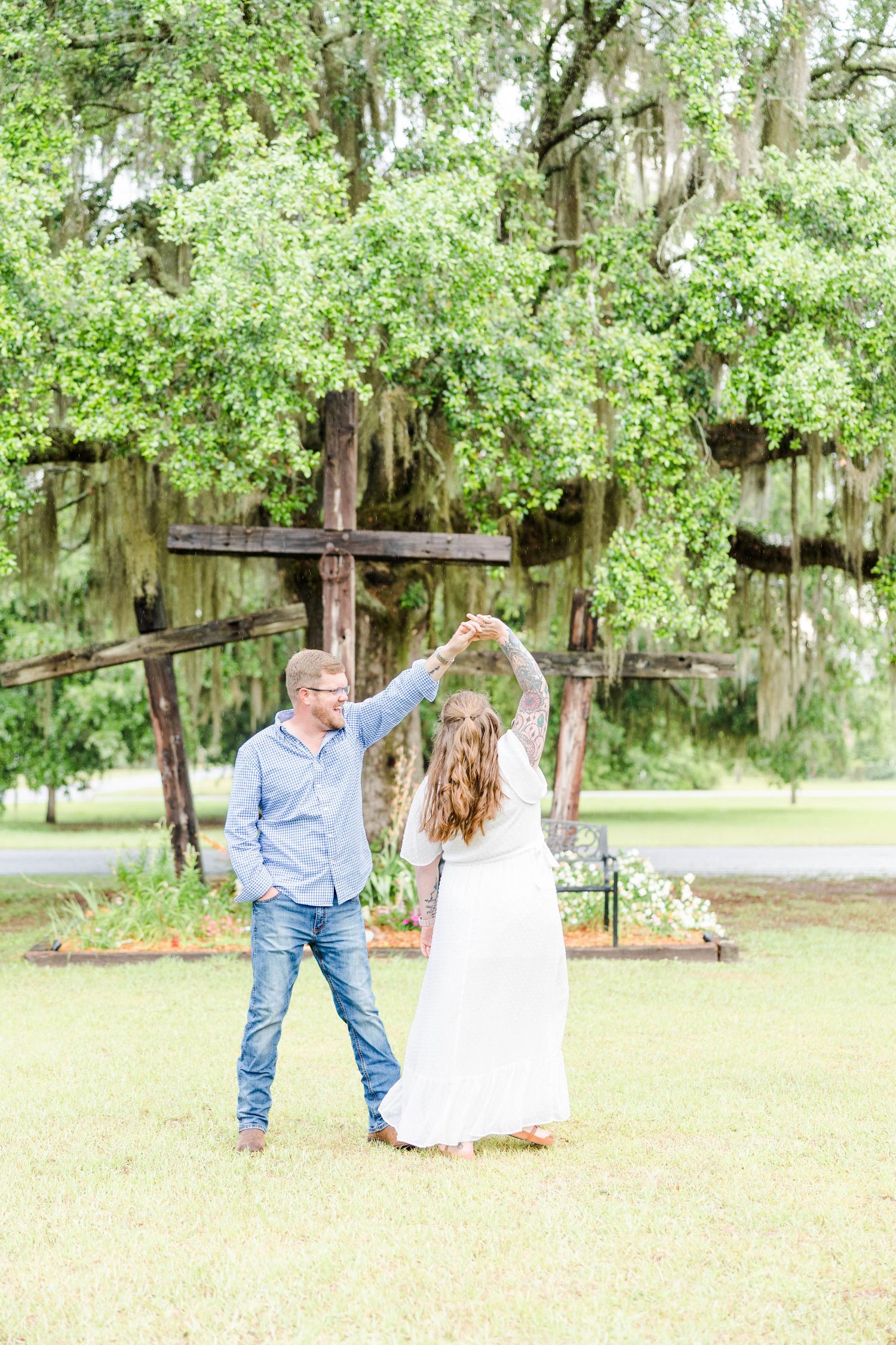 A couple dances together on grass near a rustic wooden cross draped with Spanish moss.