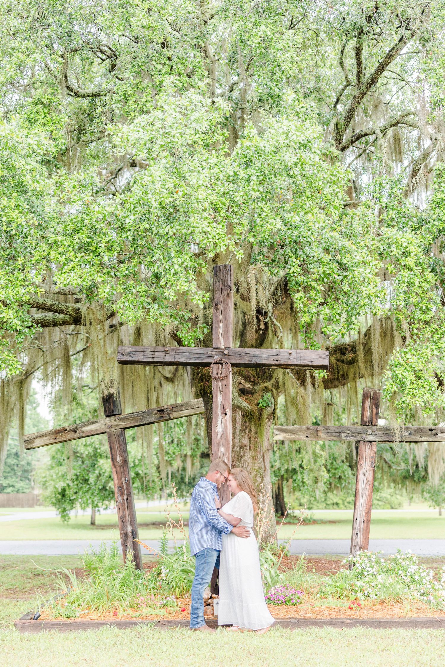 A couple shares a romantic moment beneath a large wooden cross covered in Spanish moss.