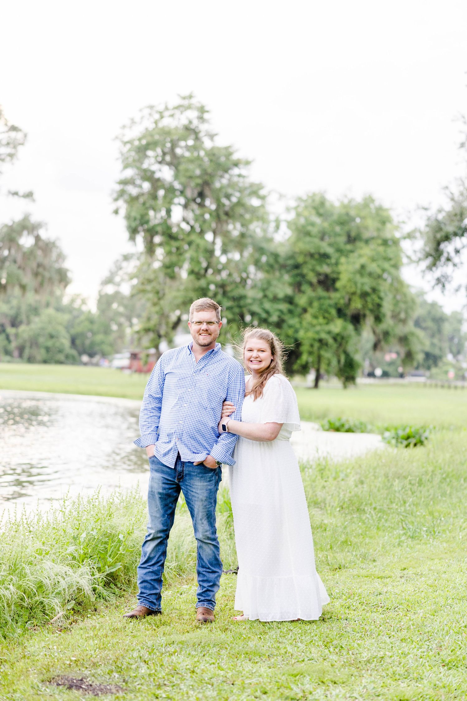 A couple poses together on grassy shoreline with Spanish moss trees in background.