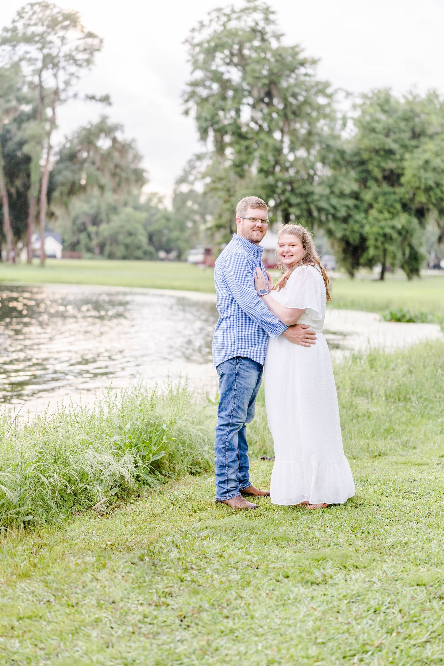 A couple embraces by a serene lake with moss-draped trees in the background.