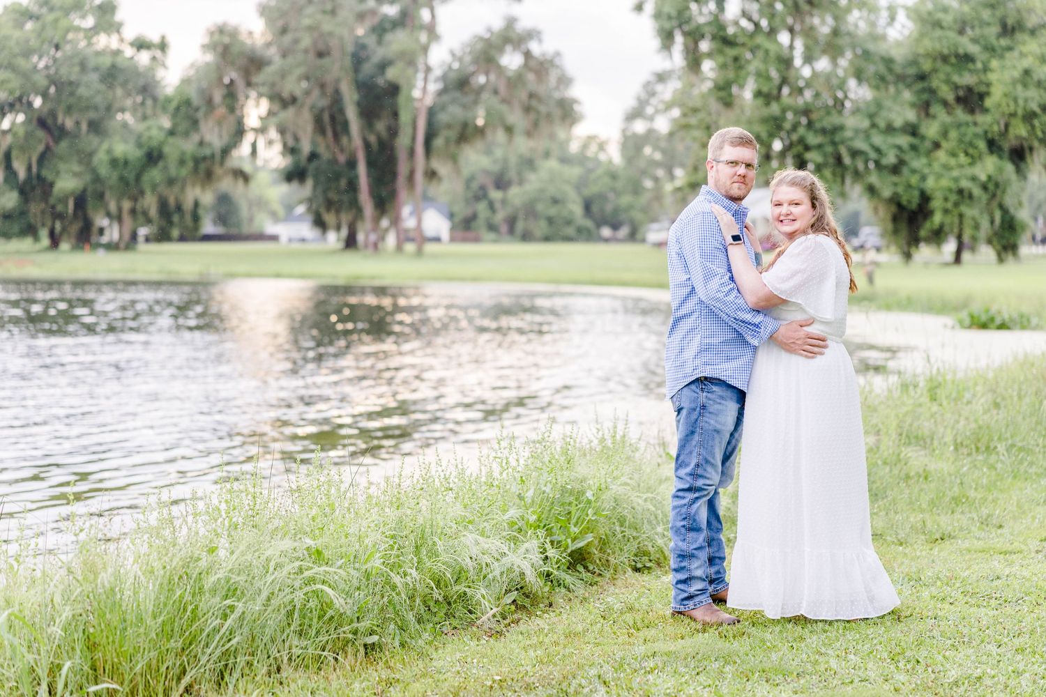 A couple shares a tender moment together on grassy lakeside in casual attire.