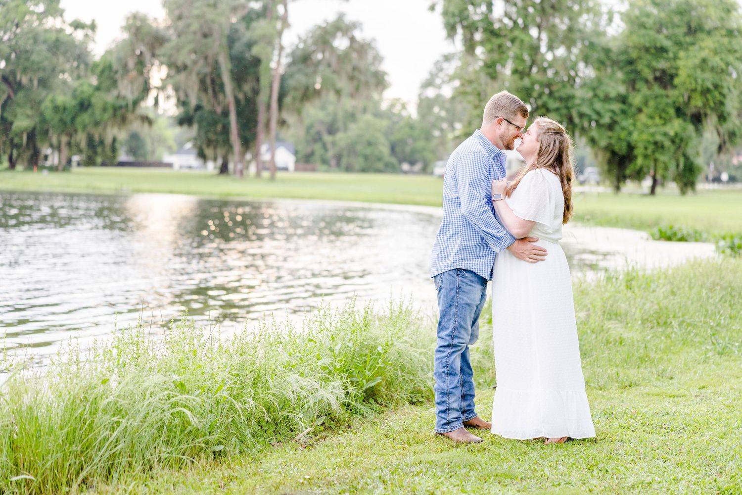 A romantic couple embraces by a serene lake surrounded by lush greenery on a bright sunny day.