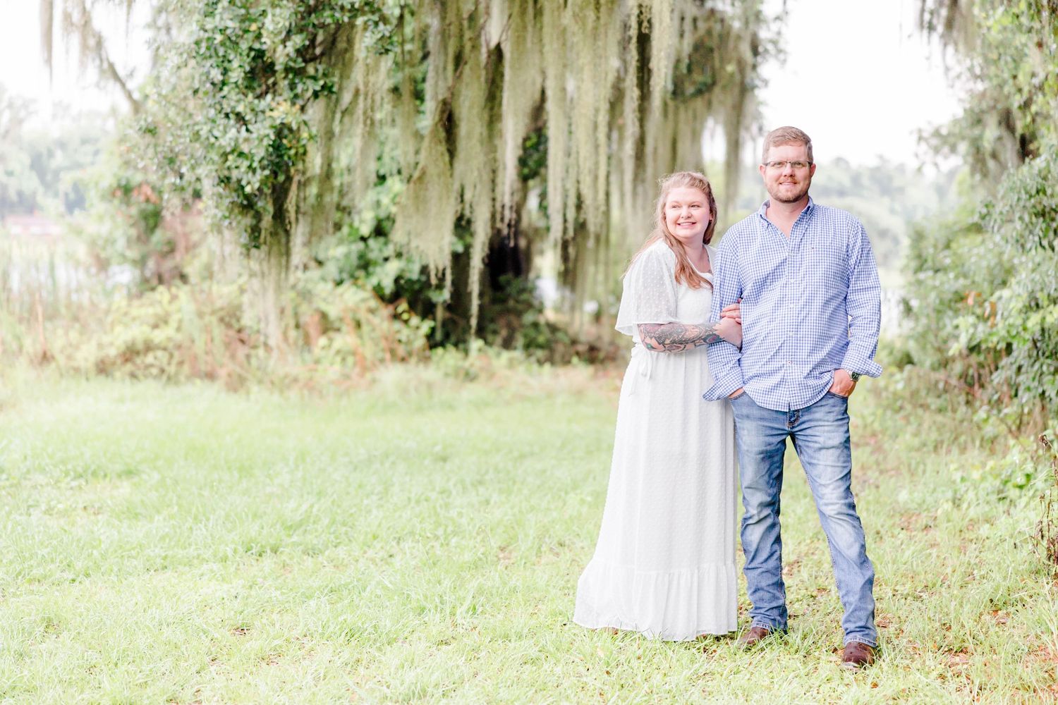A couple stands together on grass beneath Spanish moss draped trees.