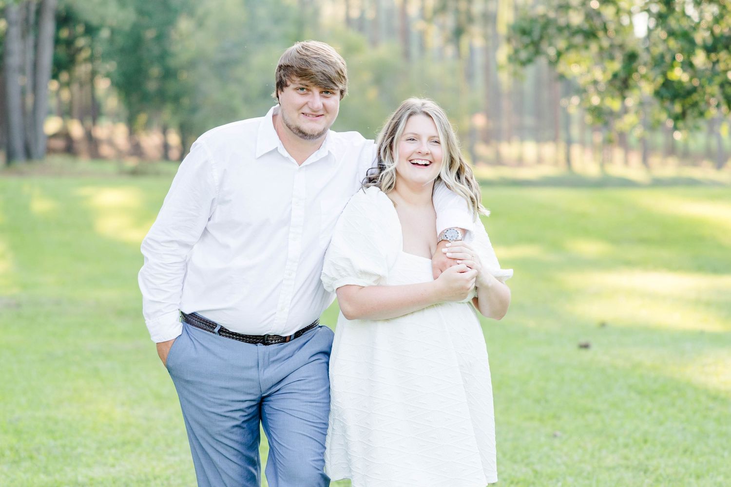 Playful couple in white and blue outfits pose for engagement photos in a grassy park.