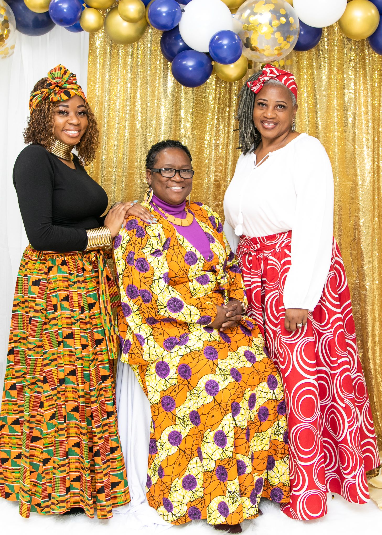 Three women in colorful African print maxi dresses posing against gold backdrop with blue balloons.
