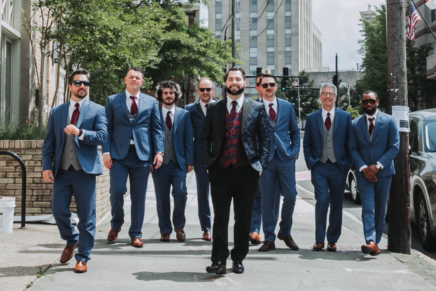 Wedding party posing on urban sidewalk with groom in center wearing black suit.