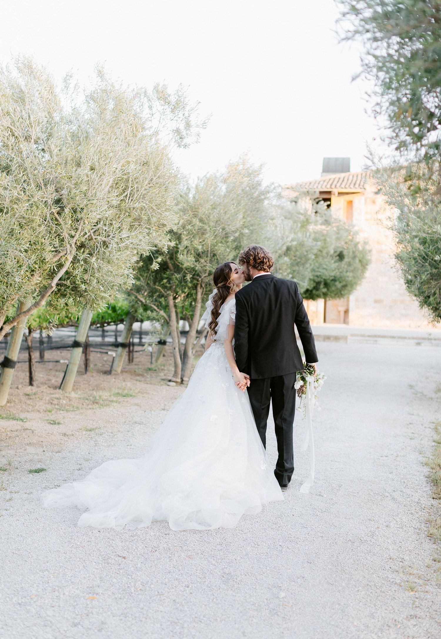 A couple walking down a tree-lined path in wedding attire.