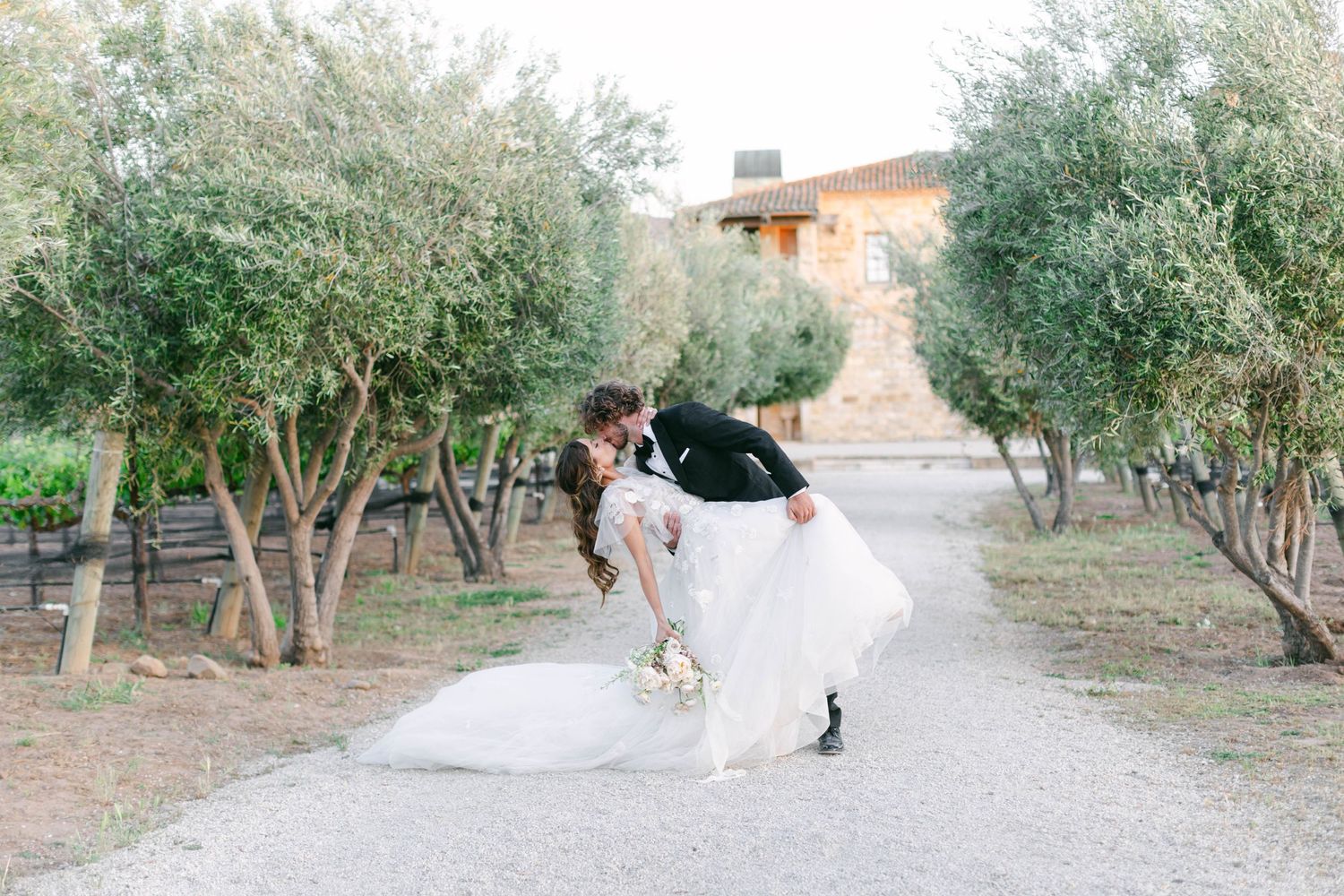 Wedding couple walks through garden pathway surrounded by trees.
