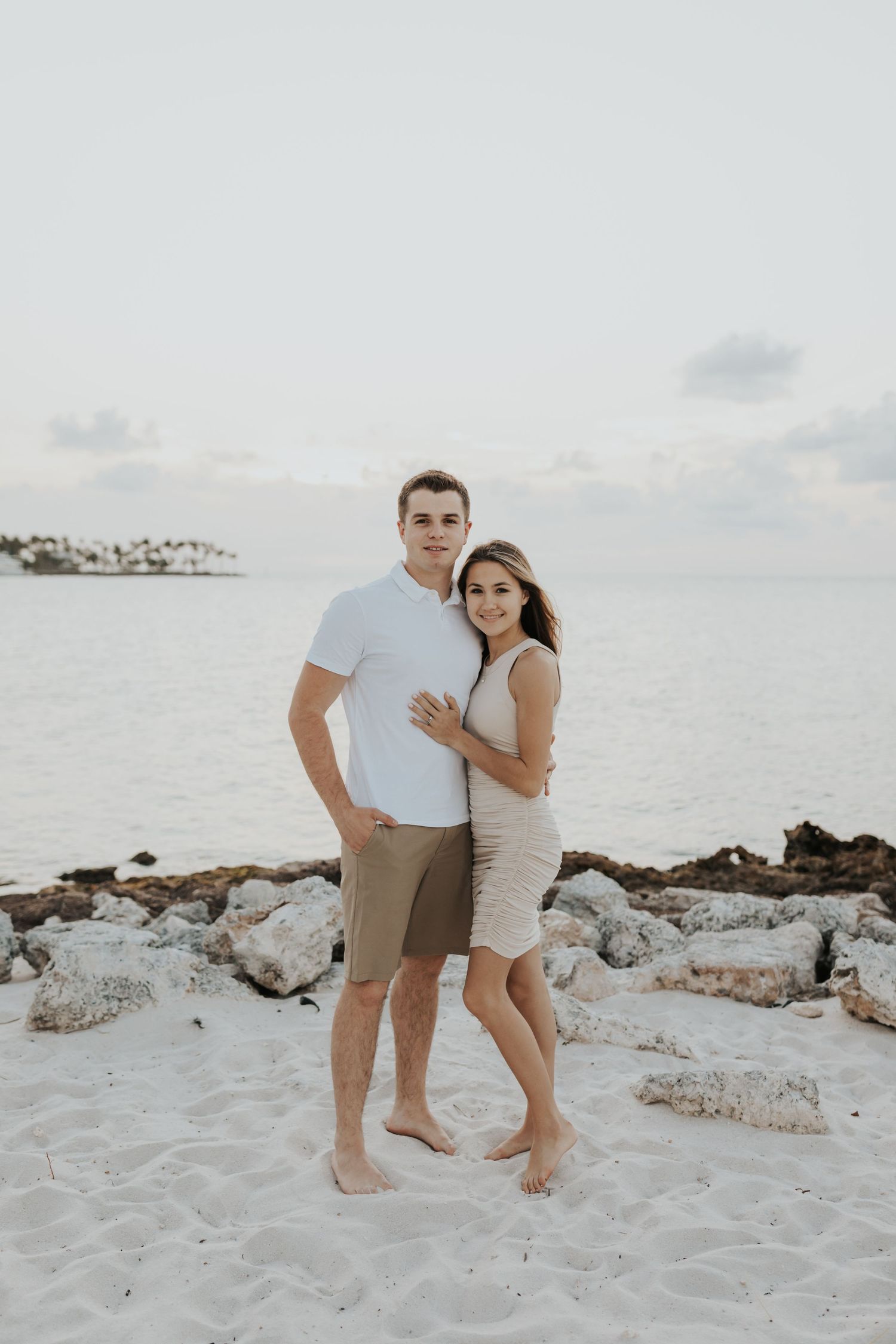 Couple poses together on white sandy beach during casual engagement photoshoot.