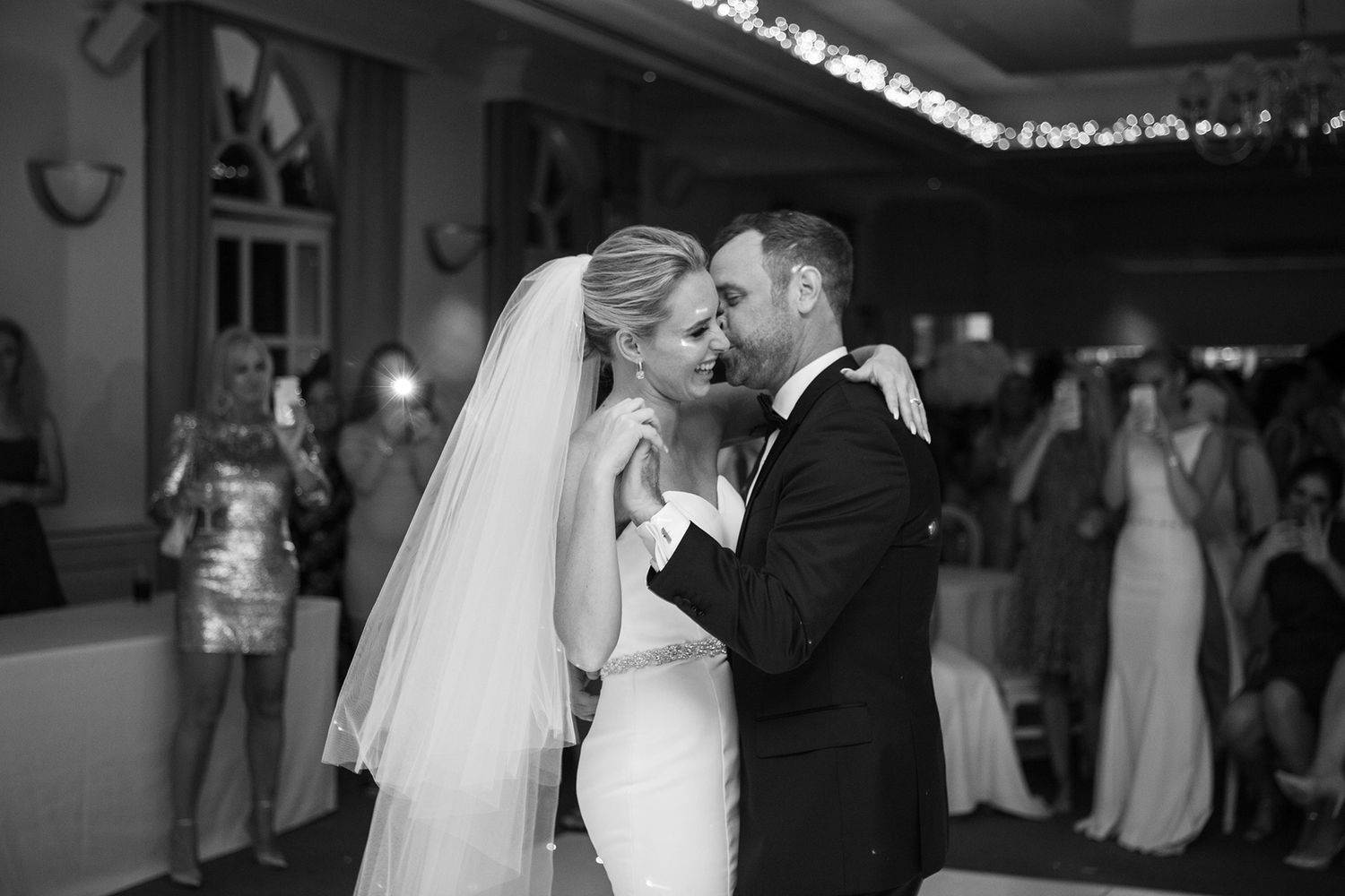 A black and white photo of a wedding reception first dance with a bride in white gown and groom in tuxedo.