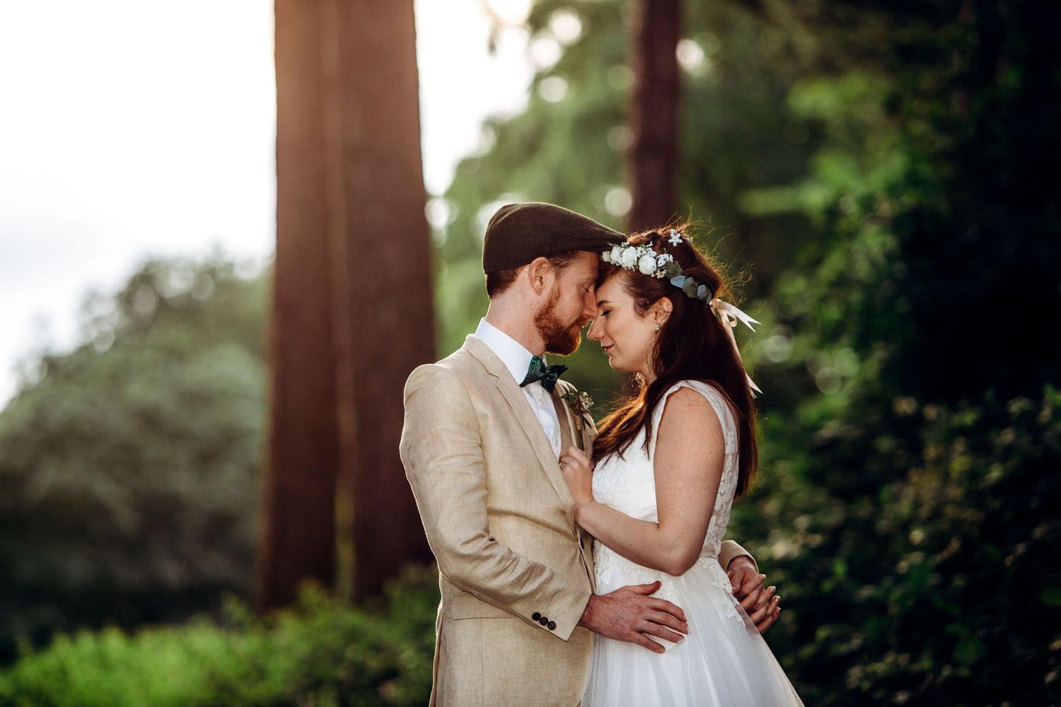 Romantic outdoor wedding portrait showing a couple in beige suit and white dress embracing in a forest setting.