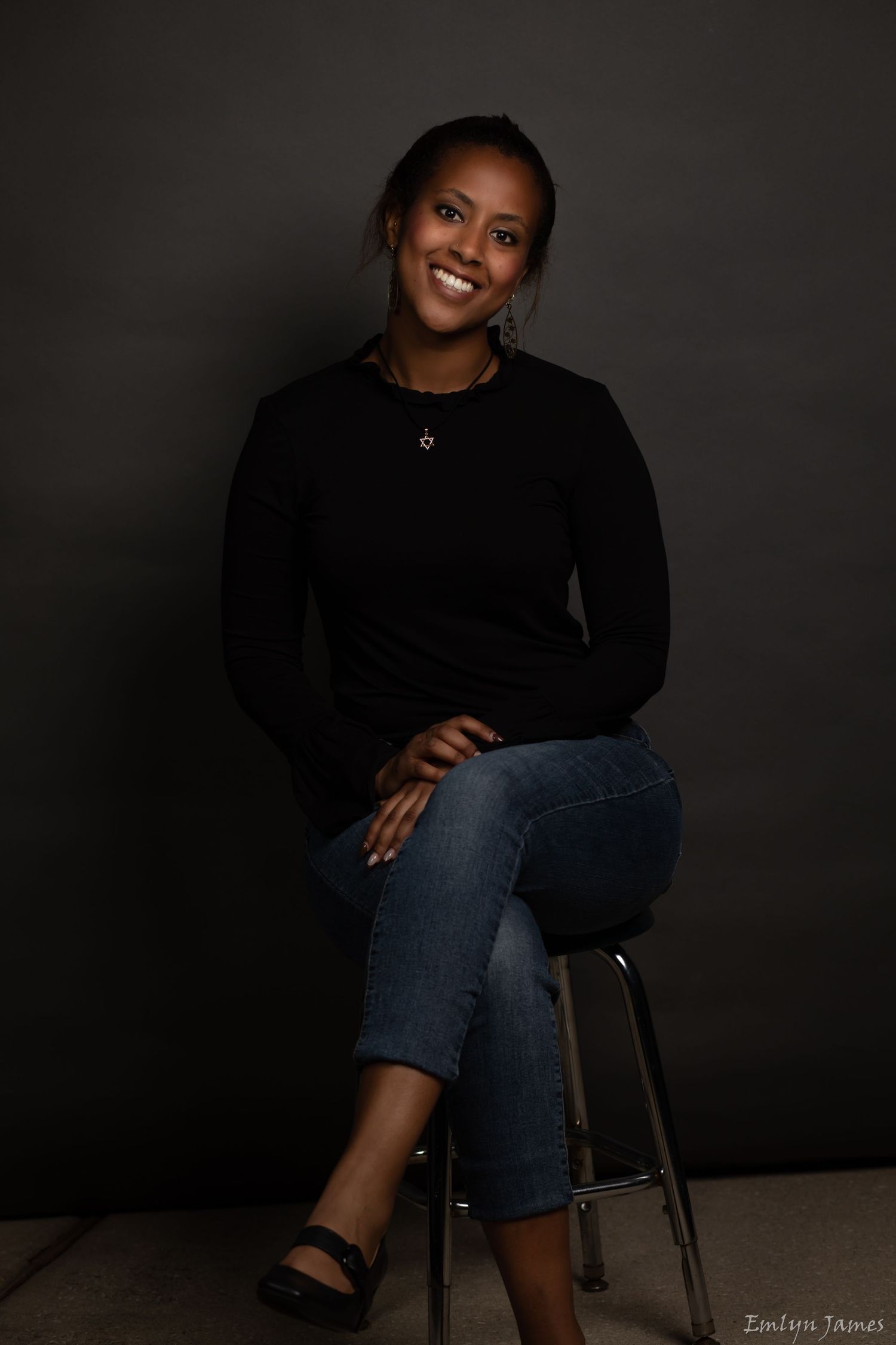Professional studio portrait of someone in black sweater and jeans sitting on stool.