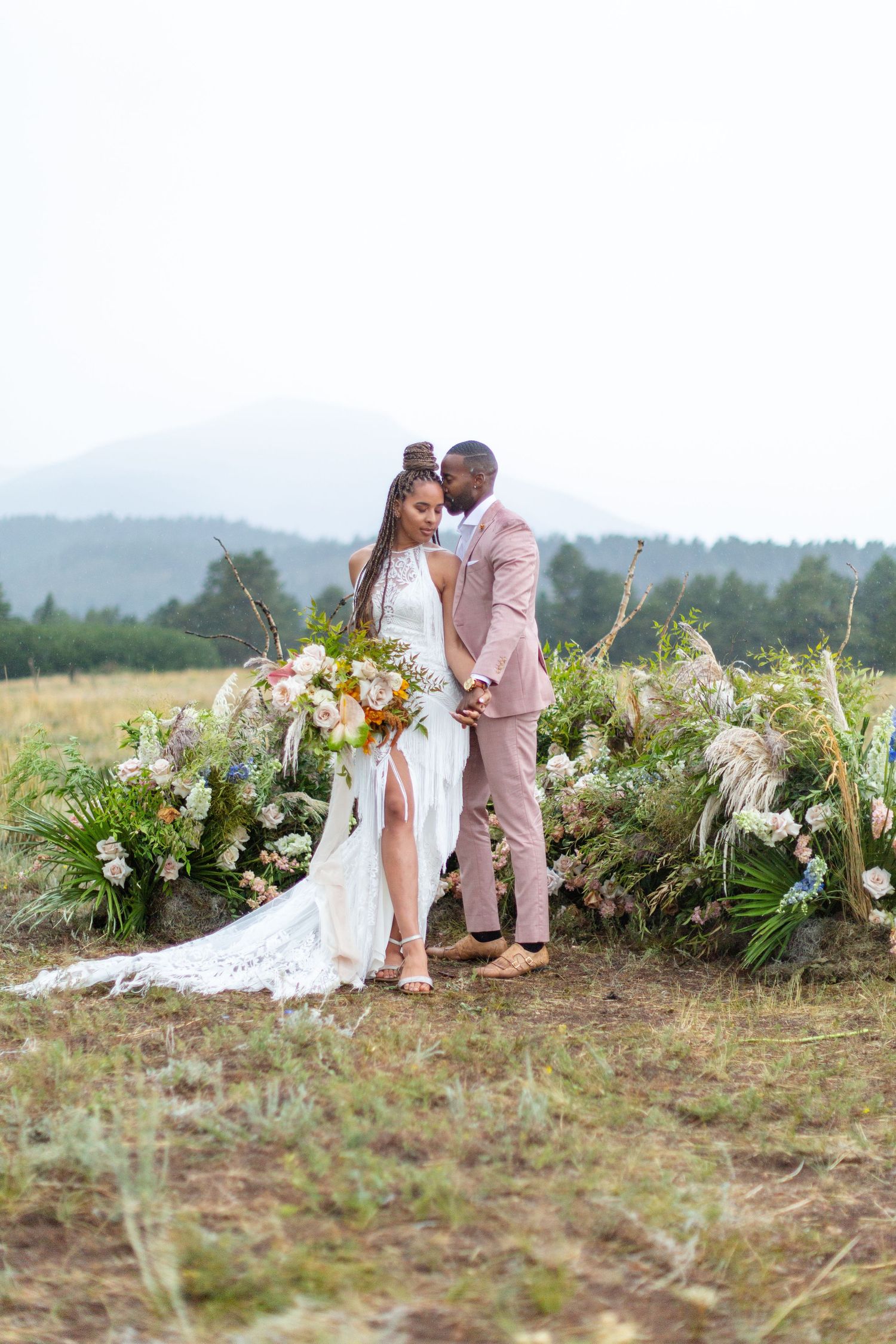 Wedding couple standing by floral arch in mountain landscape setting.
