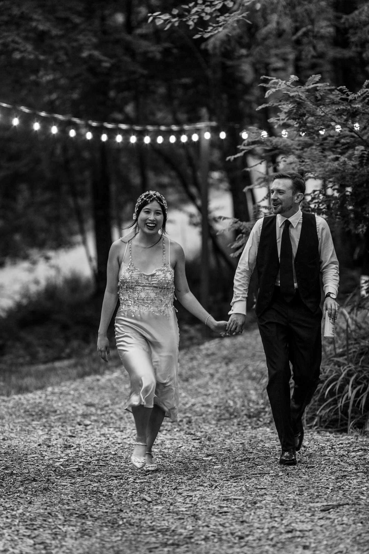 A couple walks hand in hand along a path with string lights overhead at dusk.