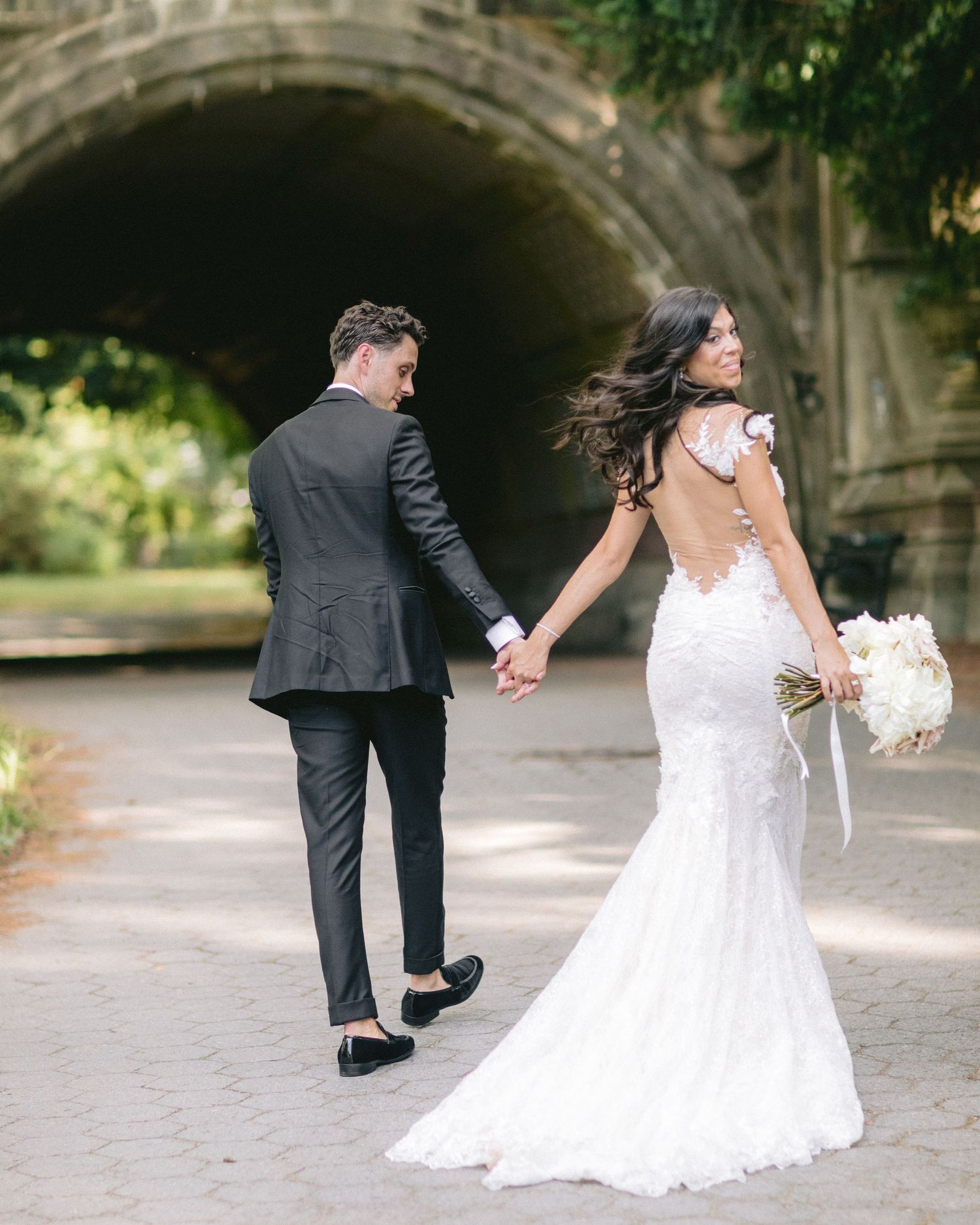 Couple walks hand in hand under stone archway with bride in backless lace gown.