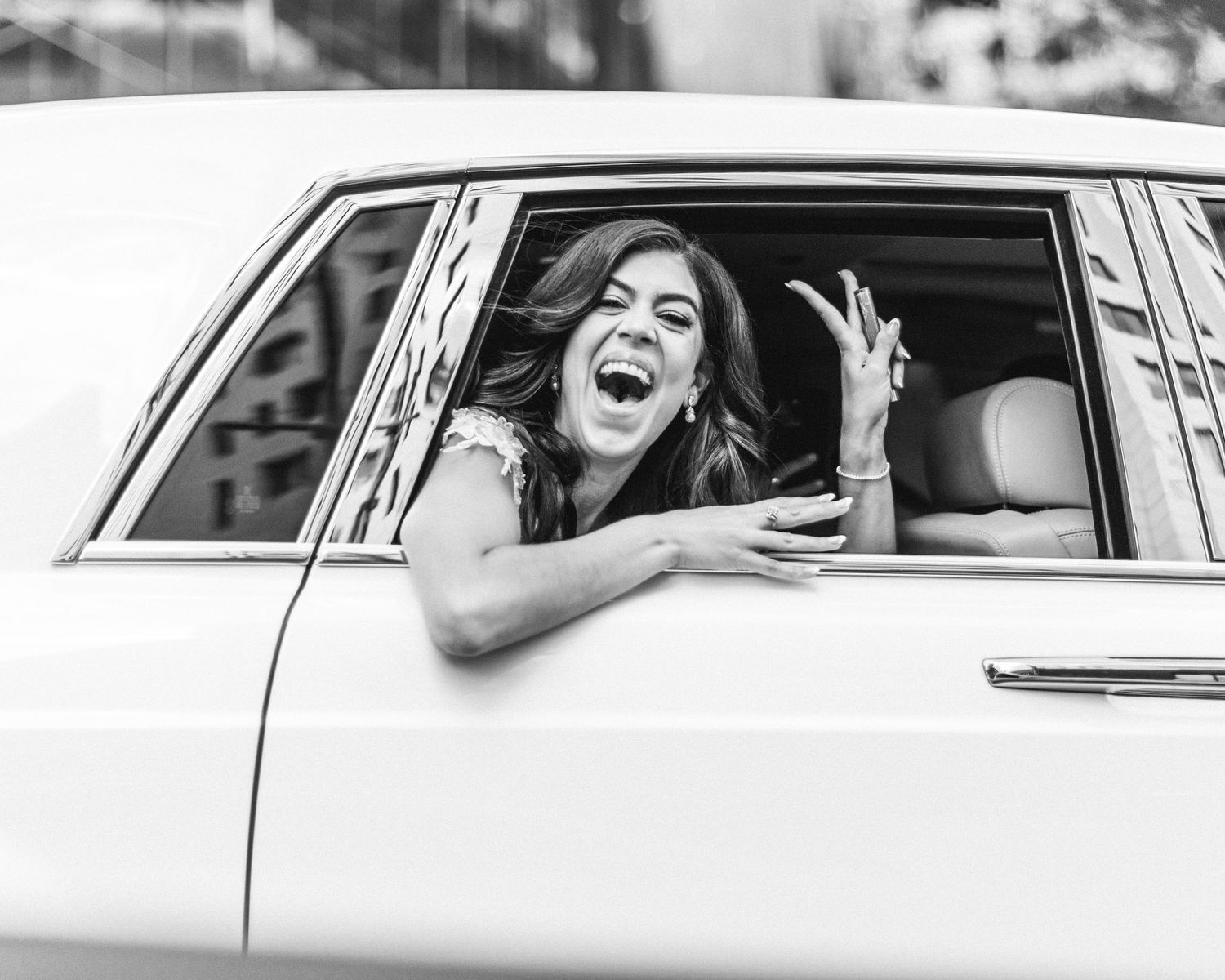 Black and white photo of bride laughing joyfully from vintage car window.