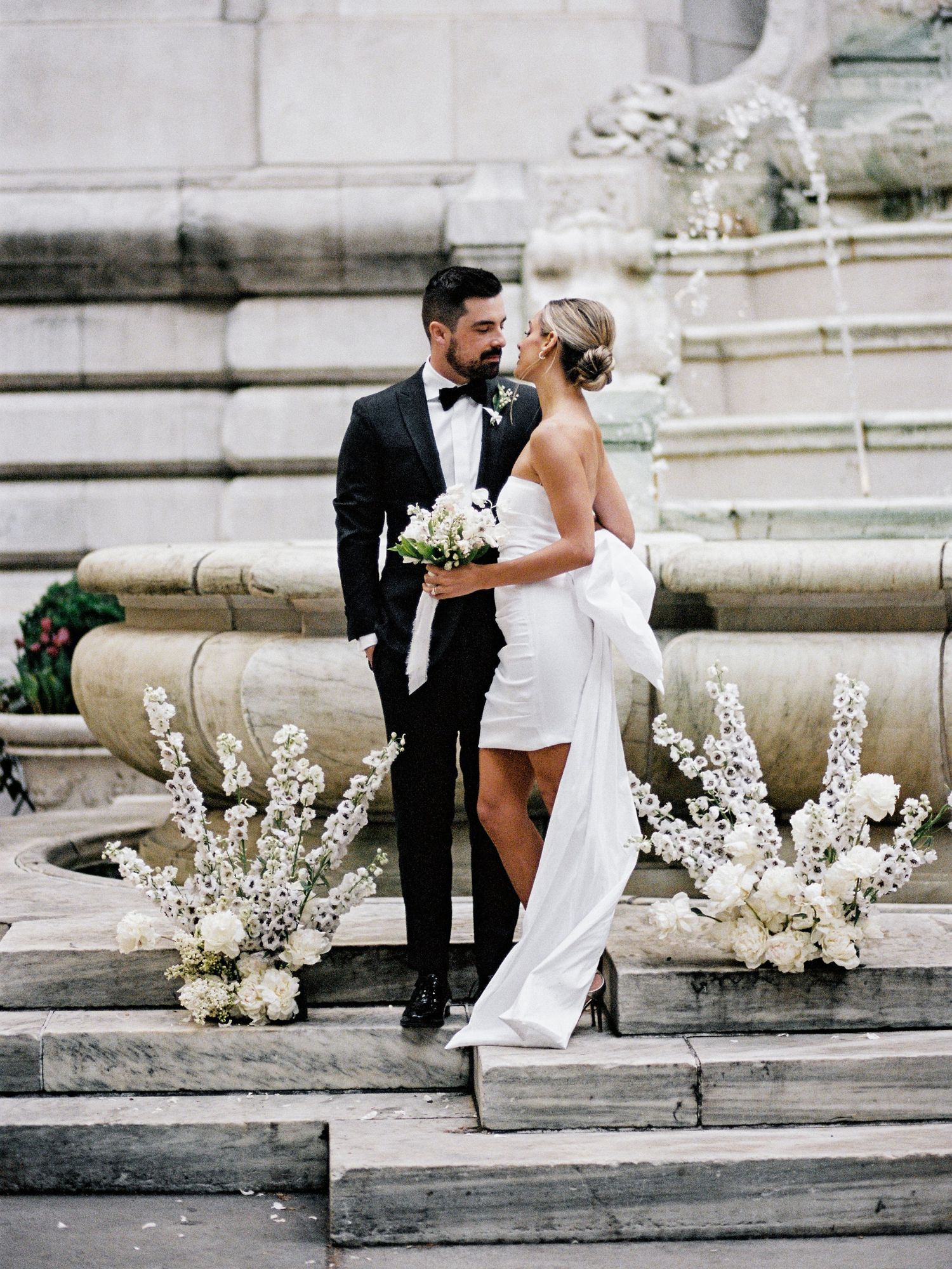Elegant bride and groom pose on stone steps decorated with white floral arrangements.