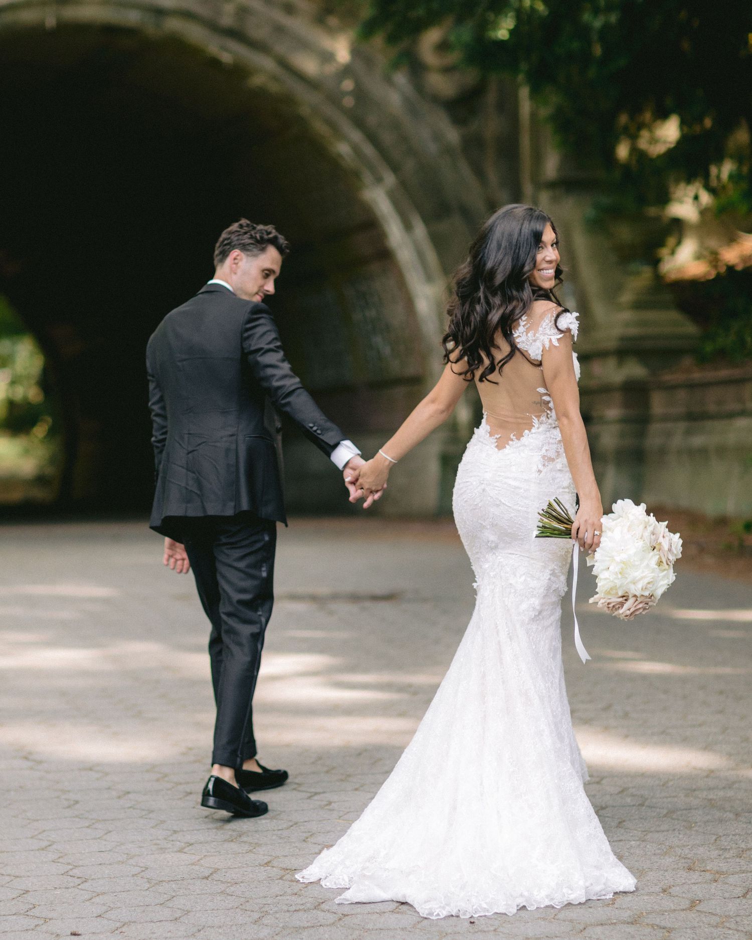 Romantic wedding photo of couple walking near stone arch with bride in lace gown holding bouquet.