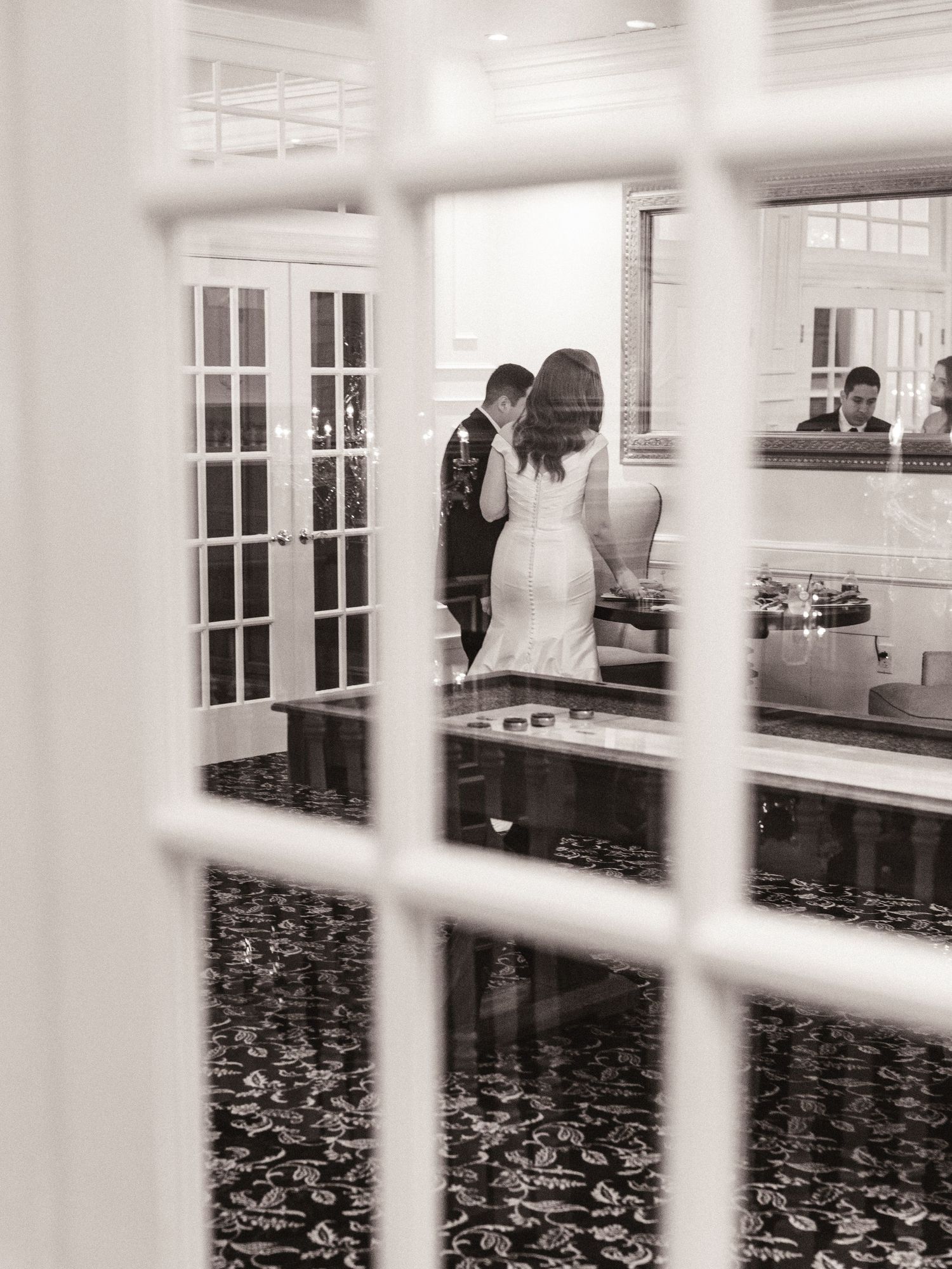 A romantic moment viewed through a window pane showing a dance in an elegant room with chandelier lights.
