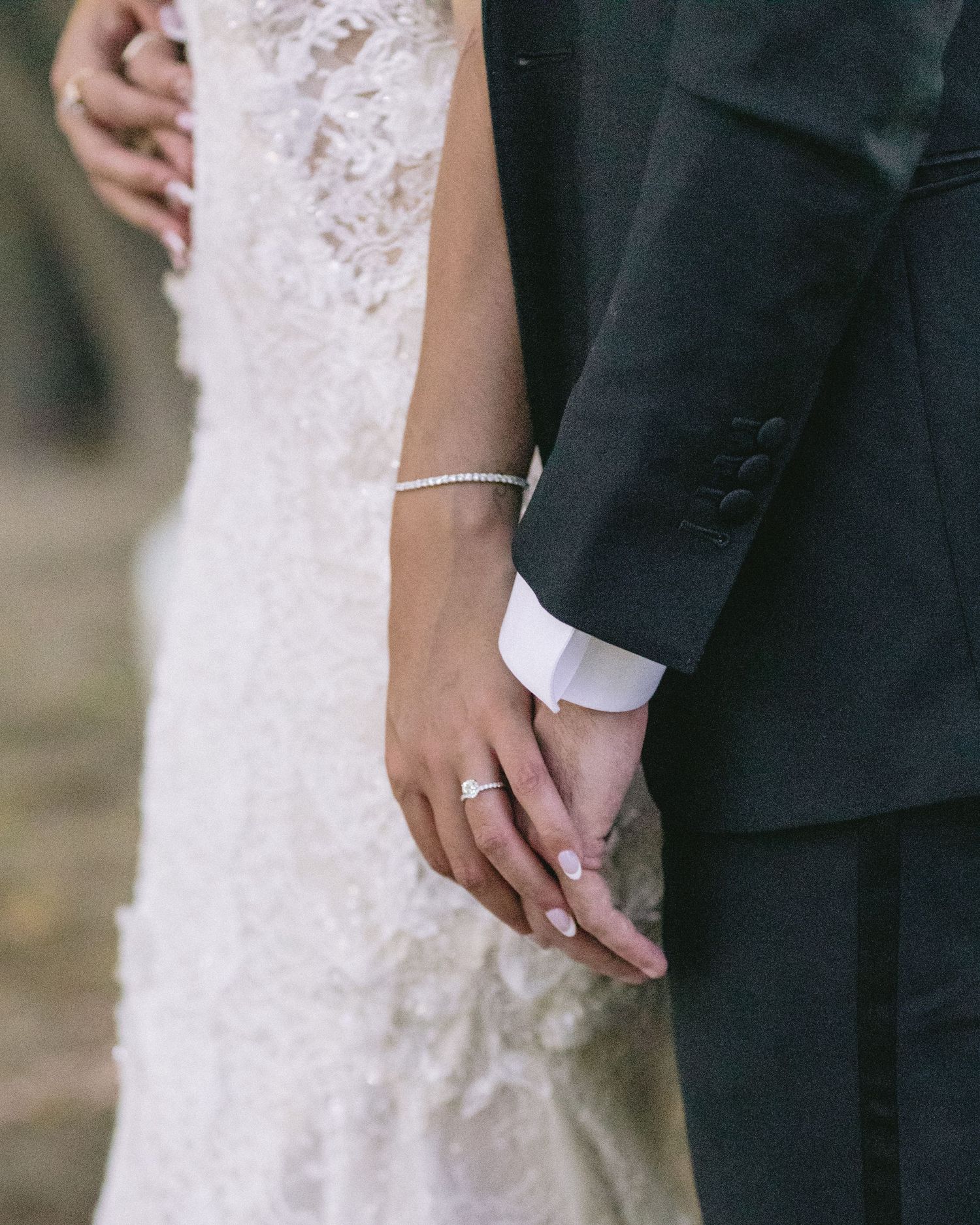 Close-up of newlyweds holding hands showing wedding rings and delicate lace dress details.