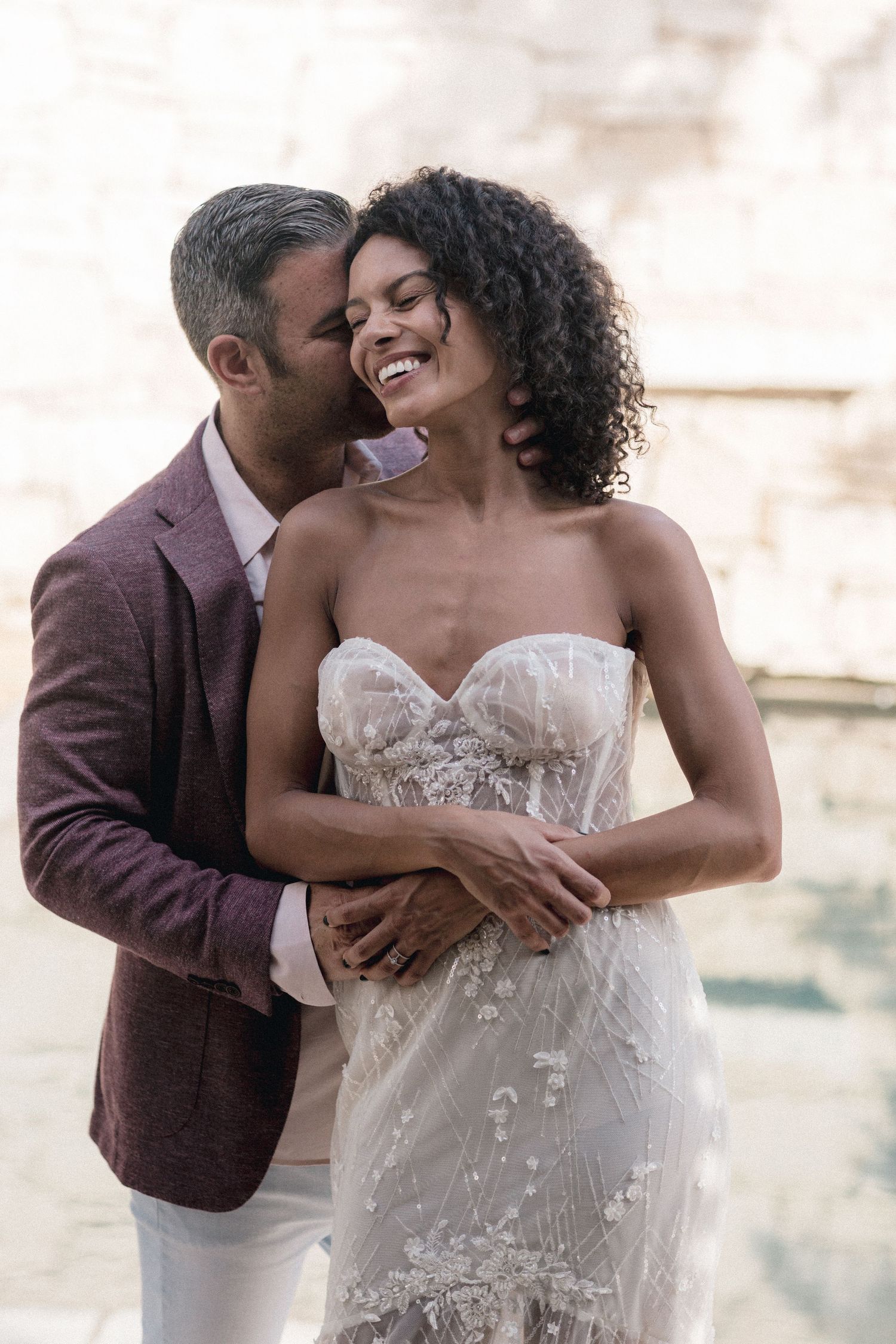 Romantic couple embracing in elegant wedding attire against light background.