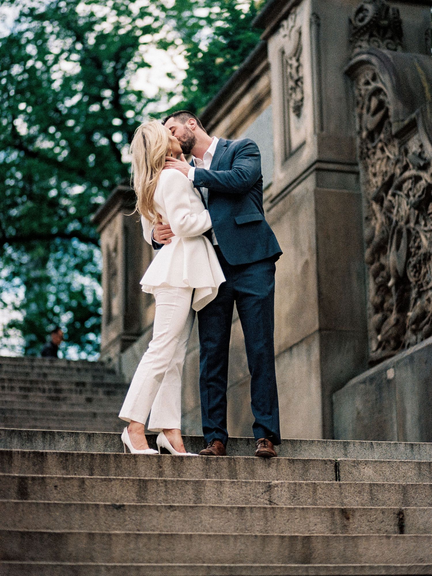 A couple embraces on stone steps against a historic architectural backdrop.