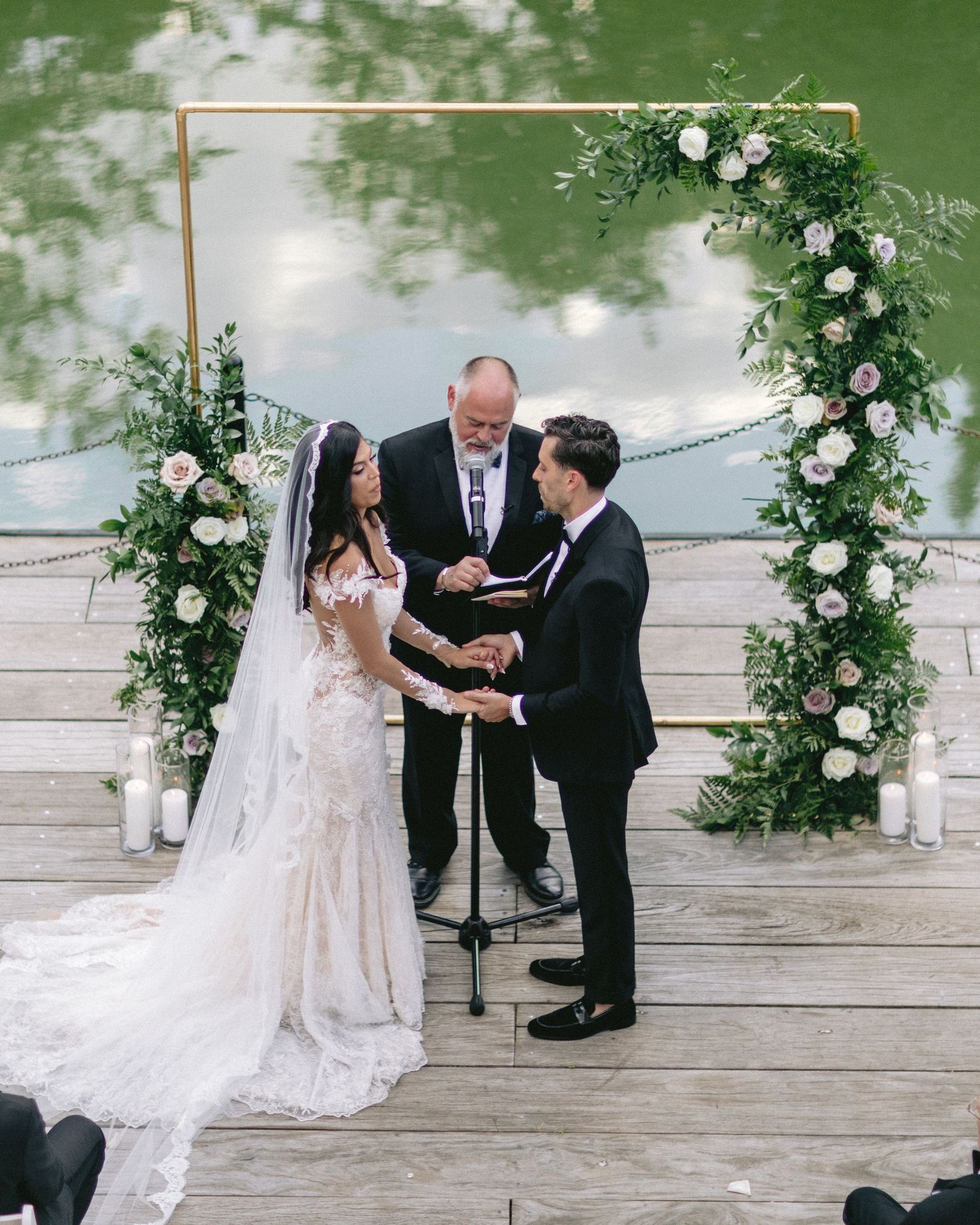 Outdoor wedding ceremony under a floral arch with white roses and greenery on a wooden deck.
