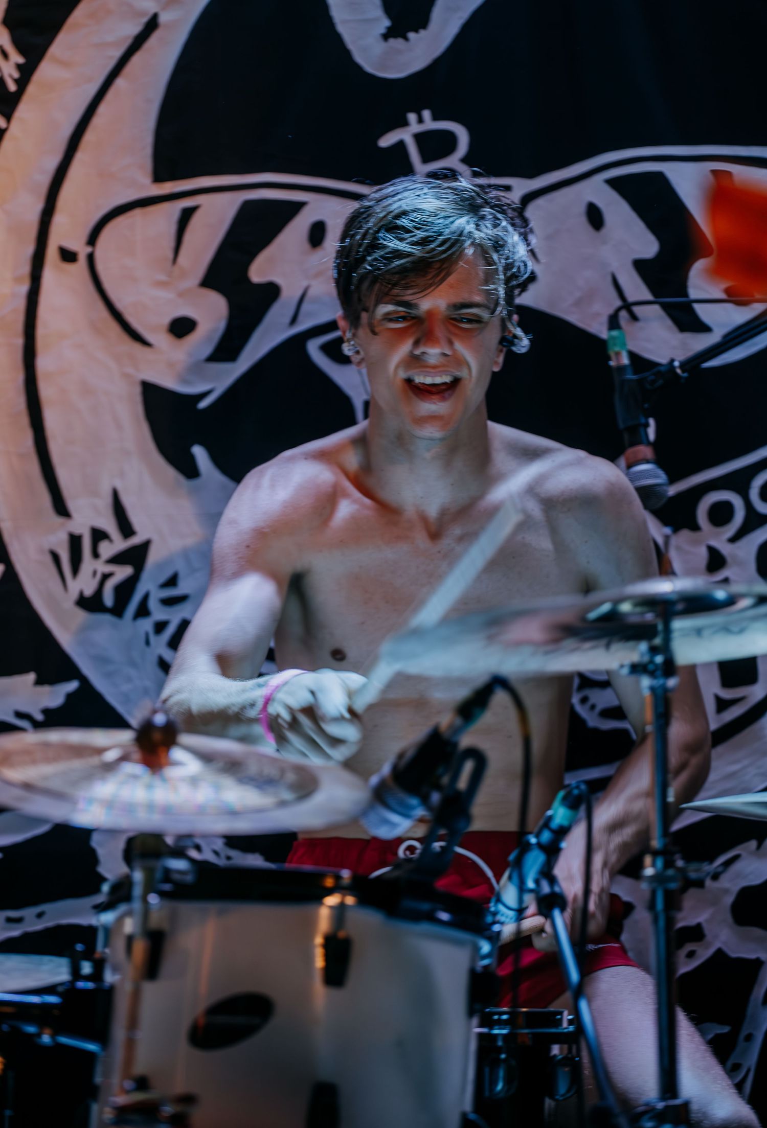 Shirtless drummer performs energetically behind drum kit with graffiti backdrop.