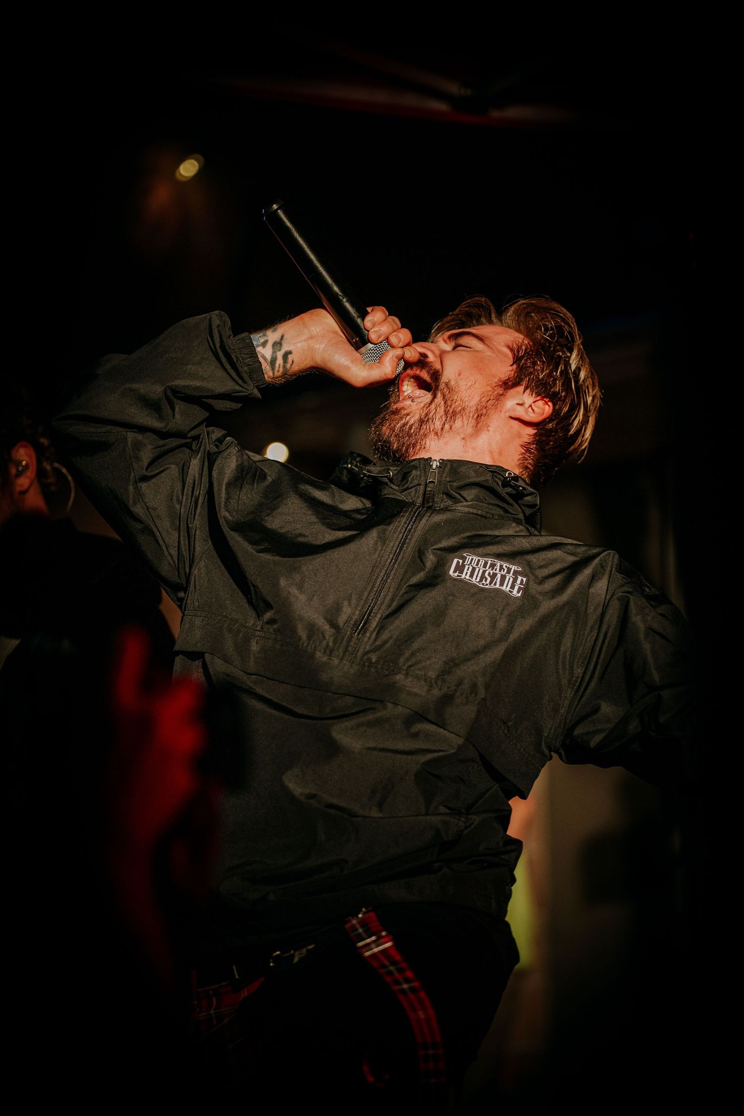 Performer in black jacket sings intensely on dark stage during concert performance.