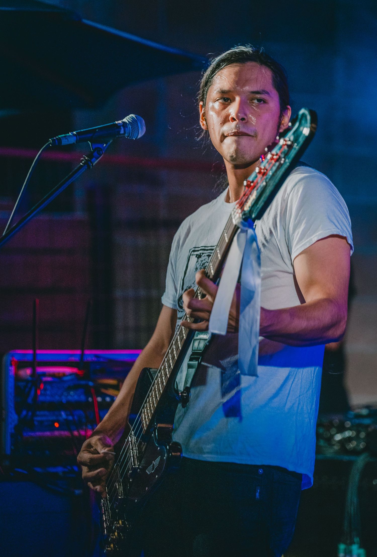 Musician in white t-shirt plays bass guitar on stage with colorful lighting in background.