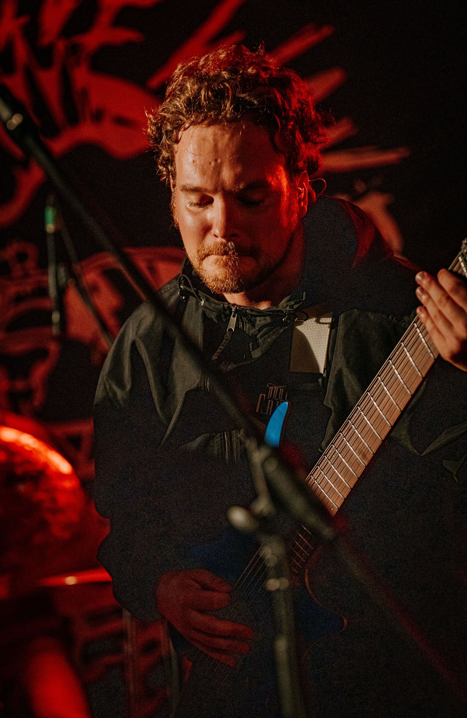 Guitarist performs in dark jacket under red stage lighting at live concert.