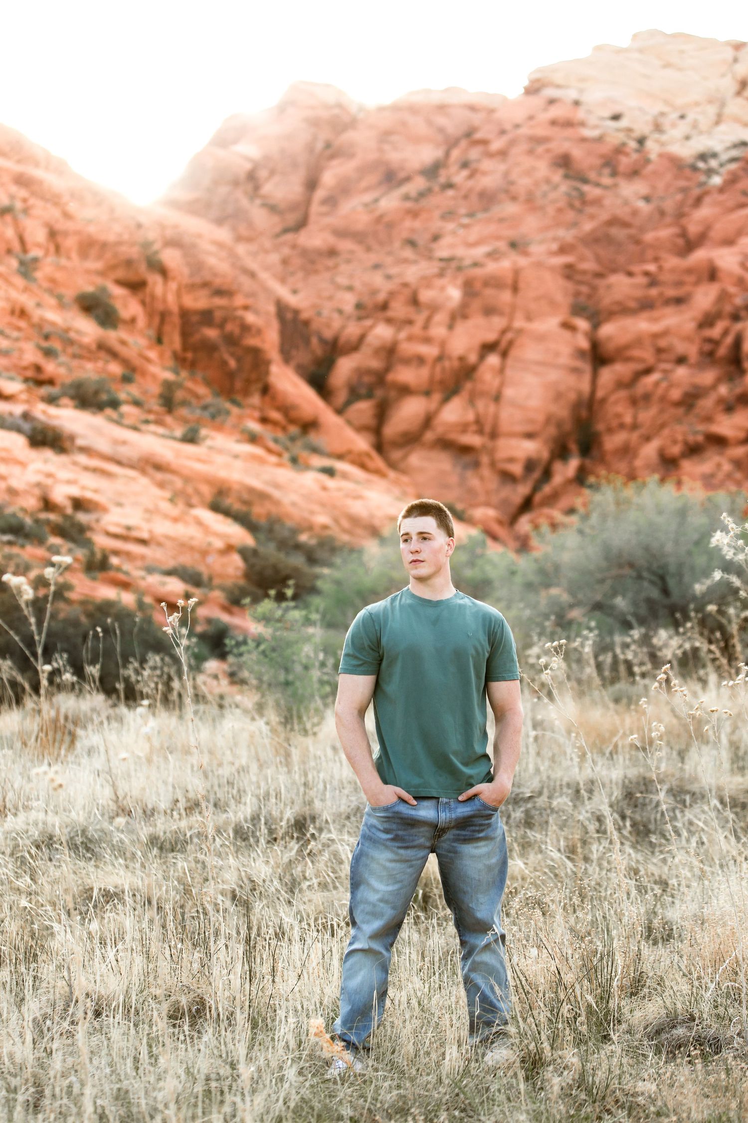 Individual in green shirt and jeans stands in desert grassland with red rock cliffs behind.