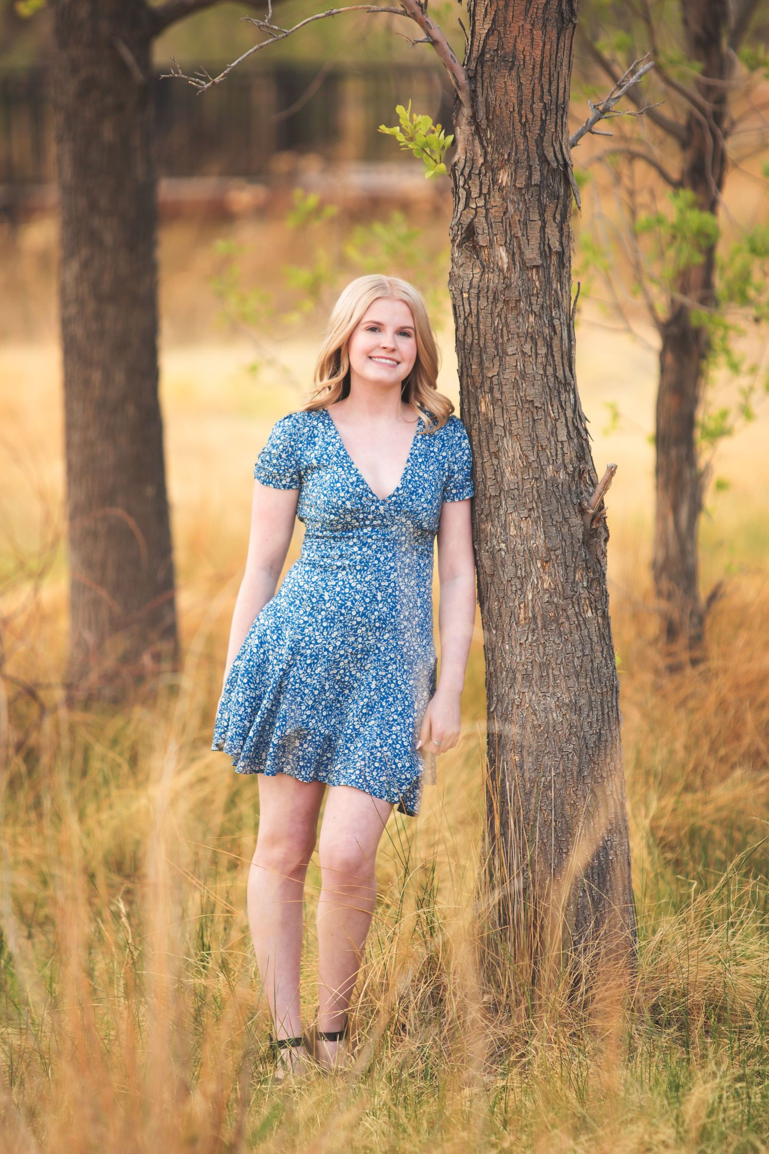 Person in blue floral dress poses playfully among trees in golden sunlight.