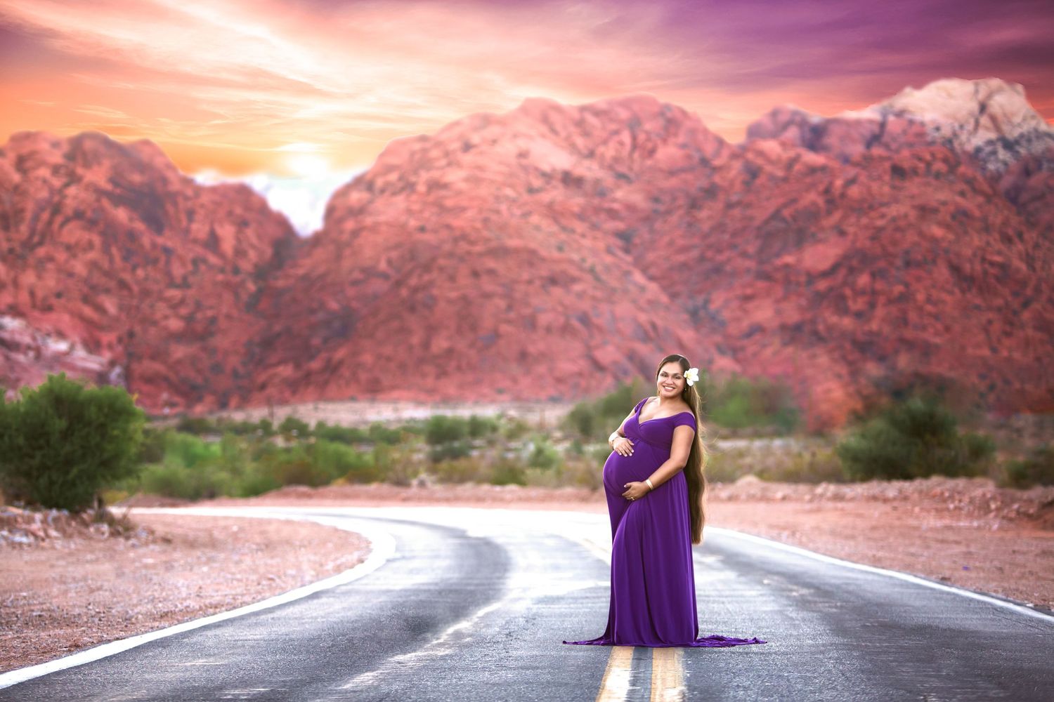 Figure in purple gown stands on desert road with dramatic red mountains in background.