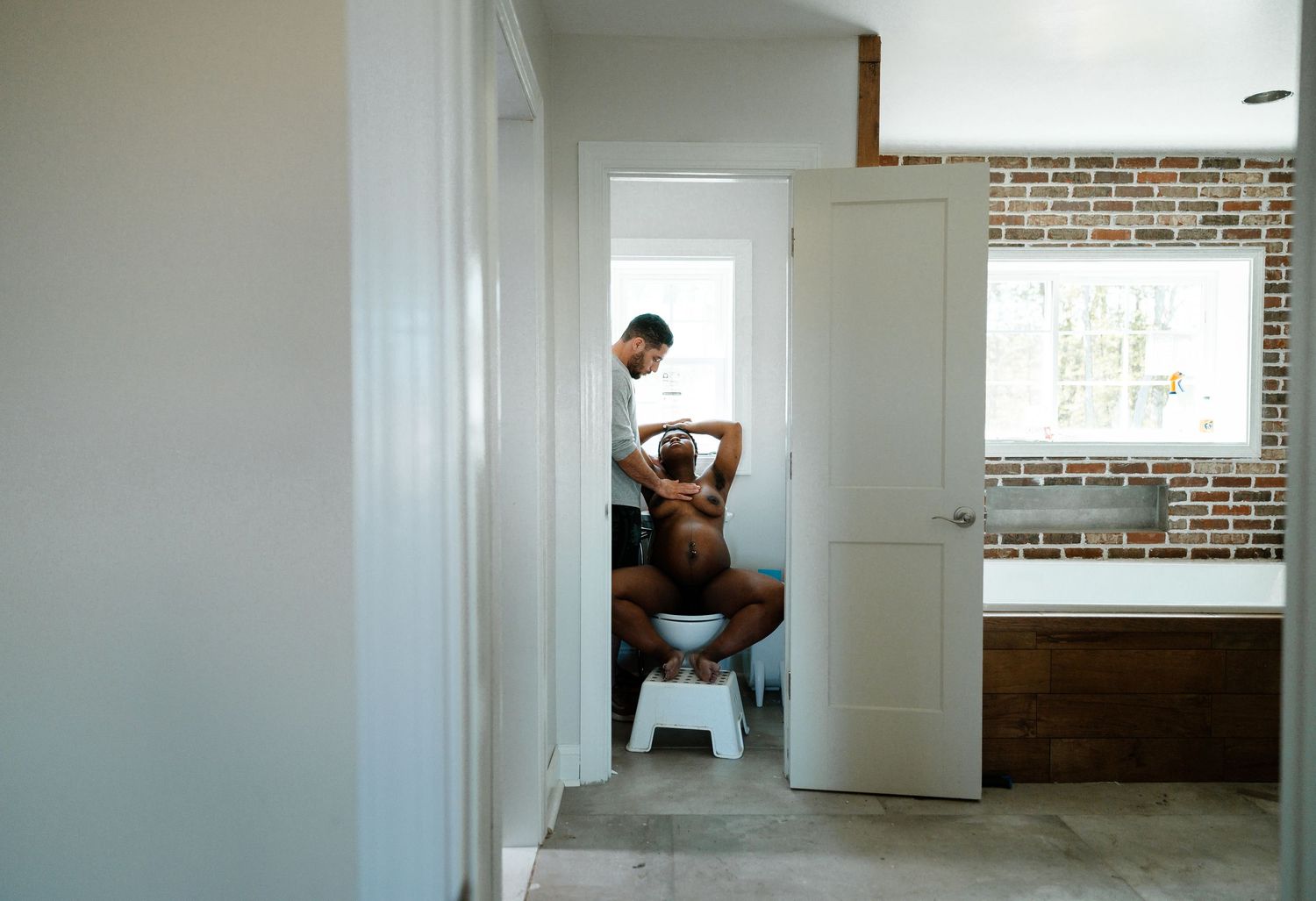 Woman Laboring At Home Sitting on a toilet With A Man Standing With His Hand On The Woman's Chest, Birth Photography