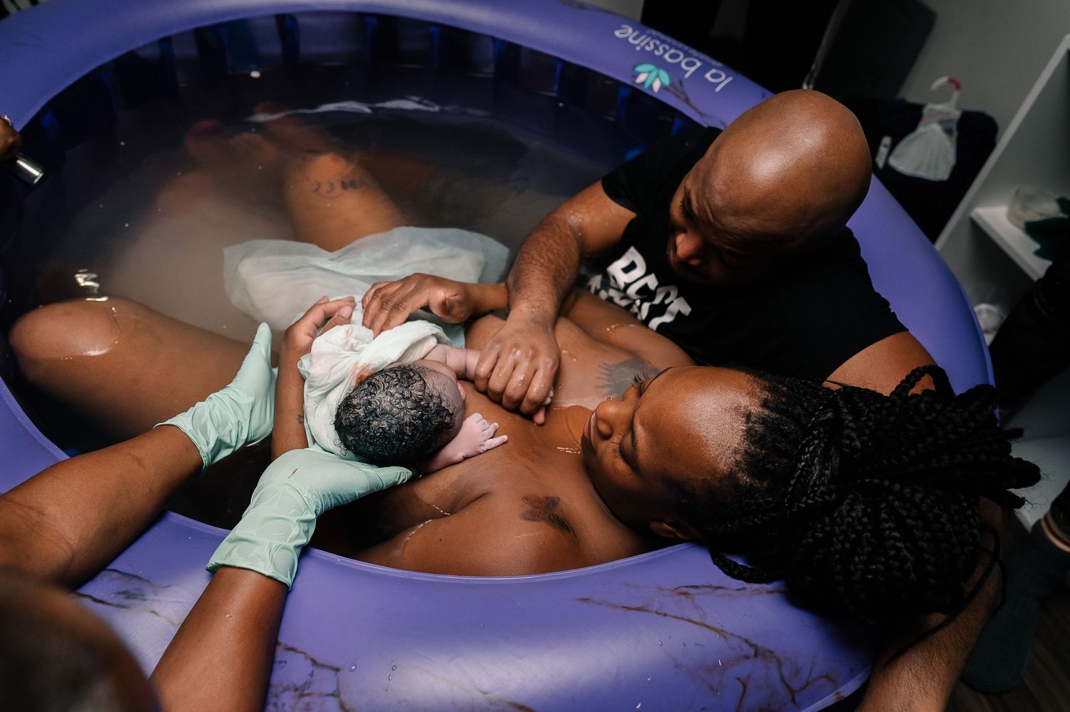 Mother and Father Sitting In Birthing Tub With Baby On Chest, Dad Holds Baby's Hand