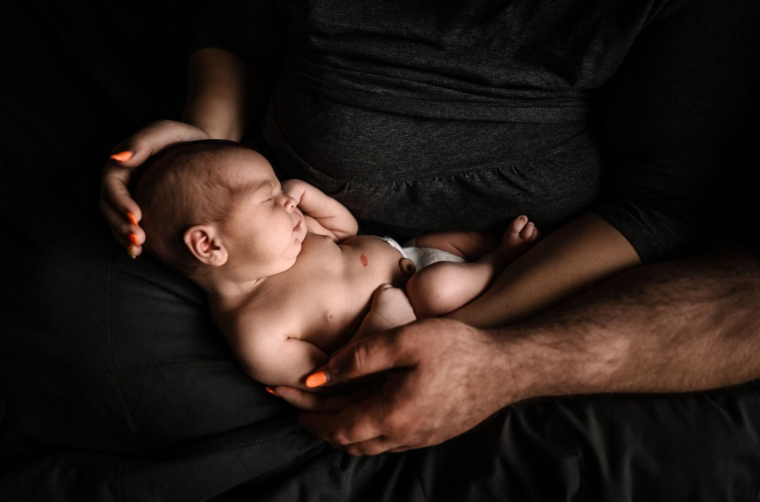 Mom and dad hold sleeping baby snuggling in woman's lap with father's hand cradling baby