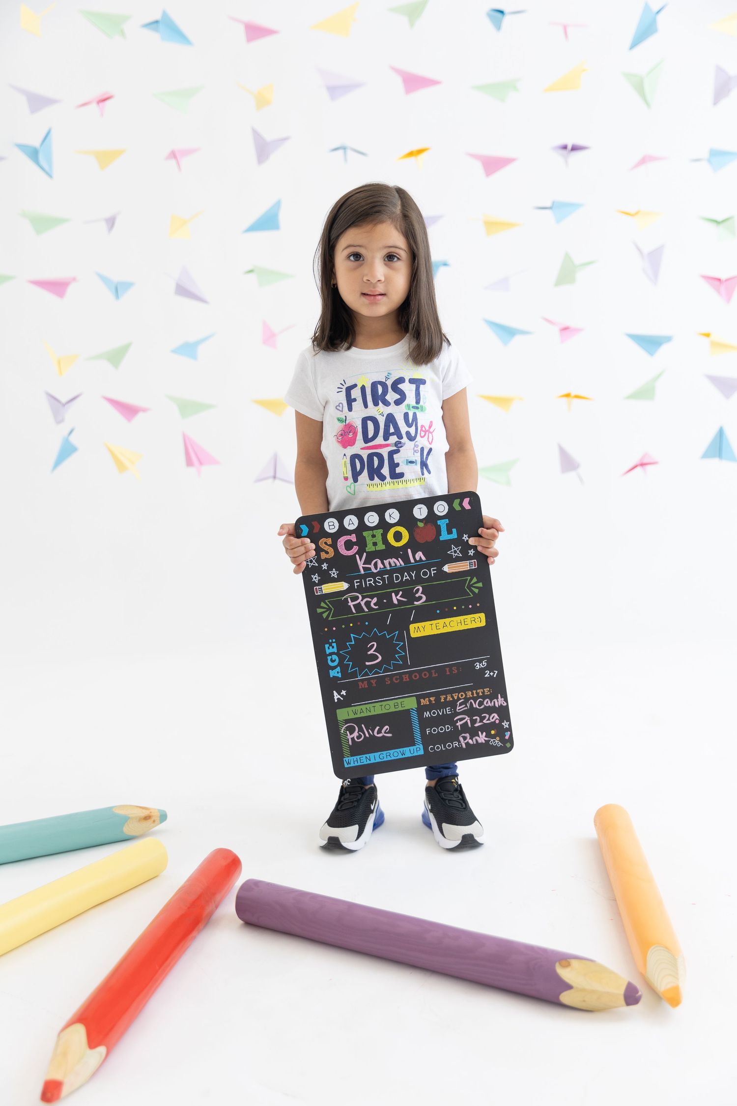 Young student holding first day of pre-k sign with decorative giant pencils on white background.