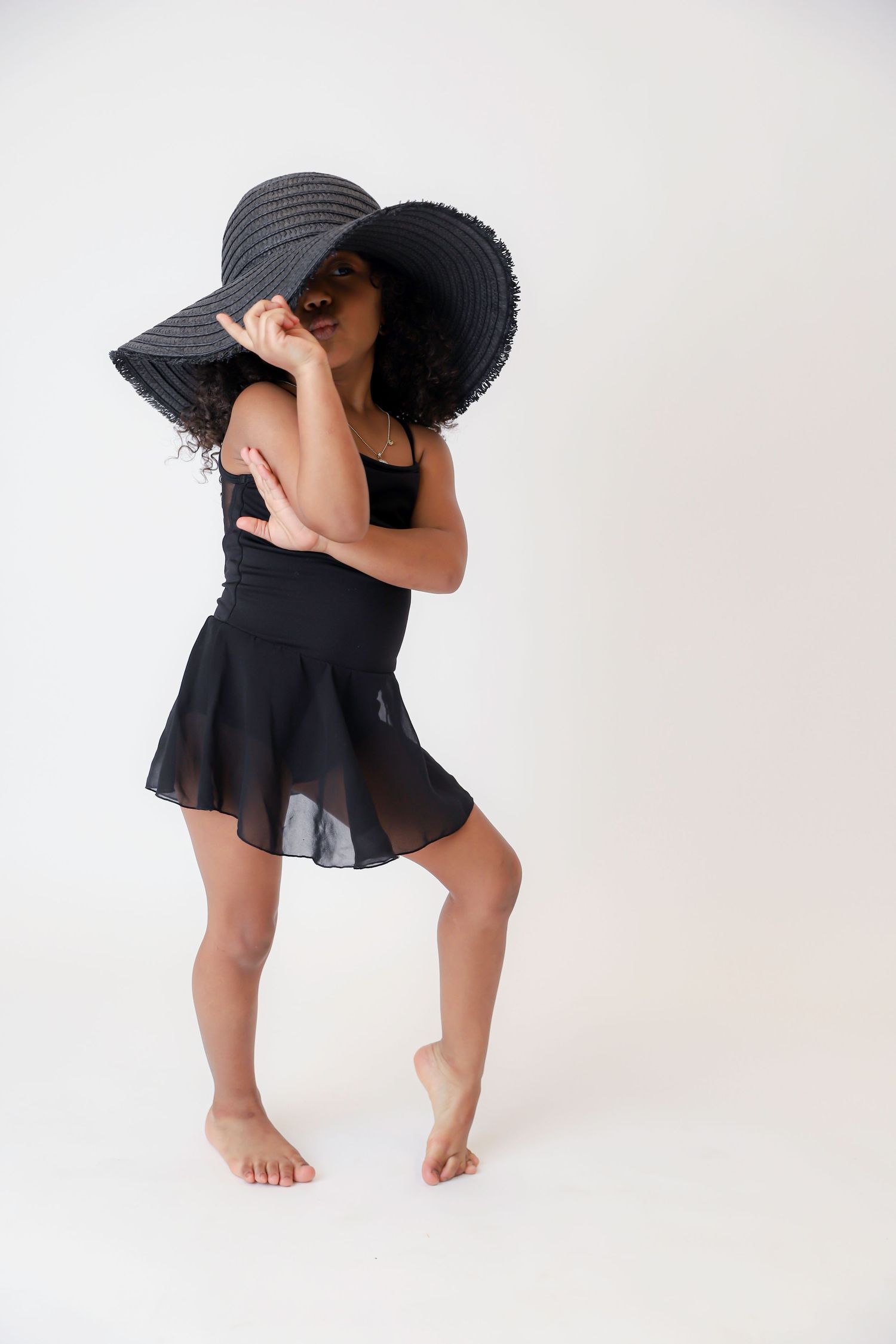 A child in black dance outfit and sun hat strikes playful pose against white background.