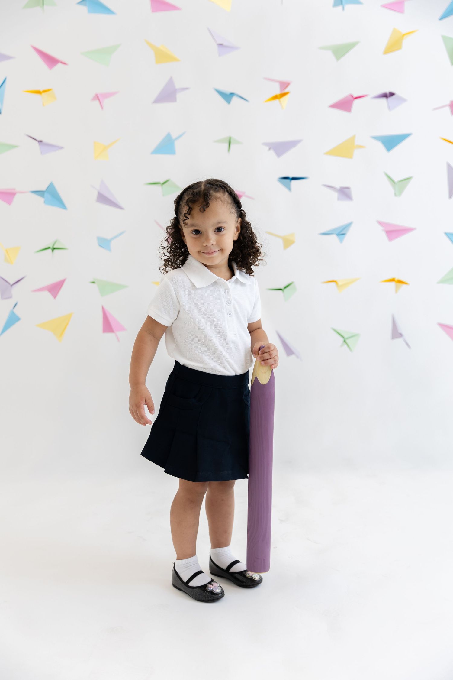 Young student in uniform holding purple pencil prop against paper airplane background.