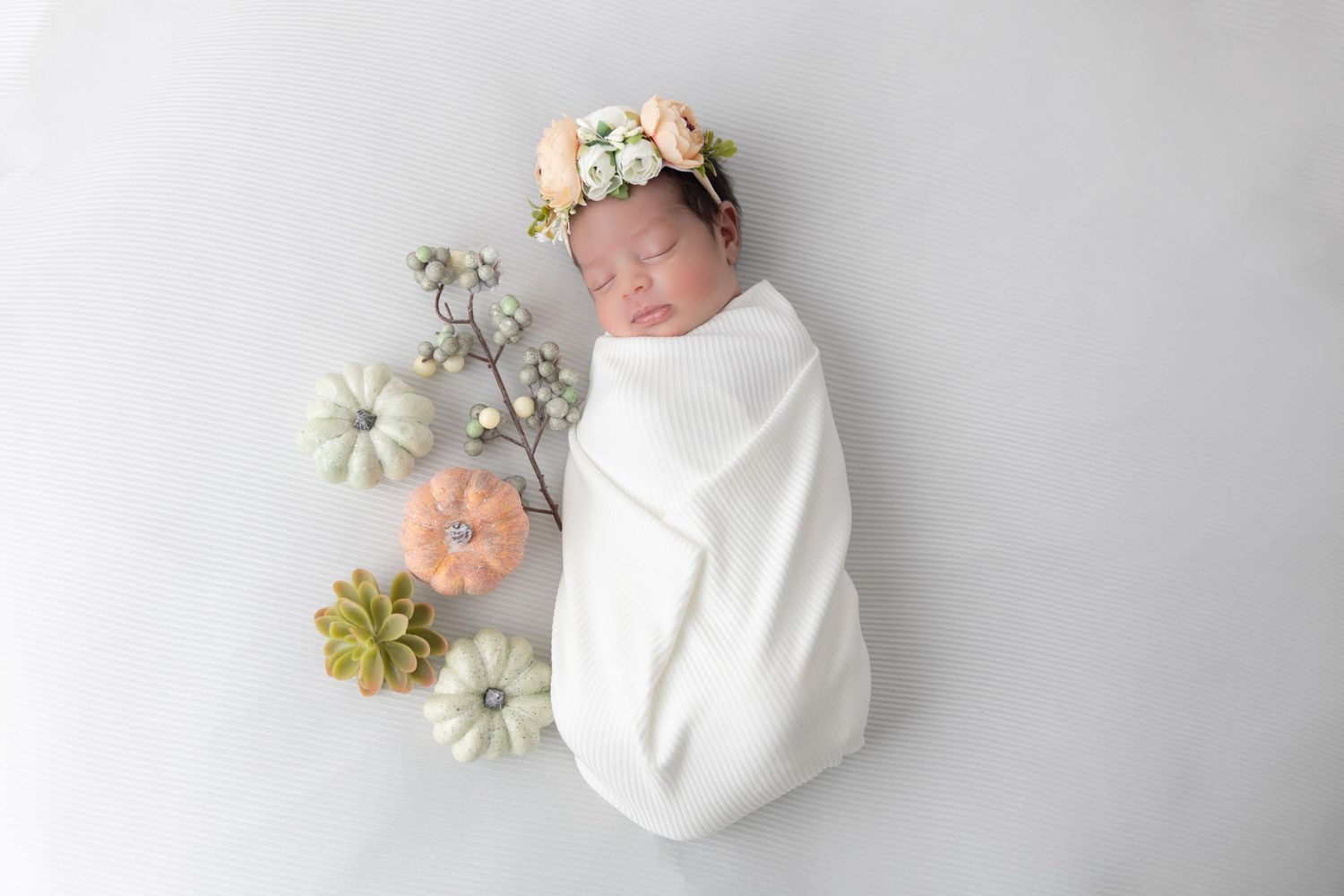 A swaddled newborn with floral accessories lies on gray background with soft pastel decorations.