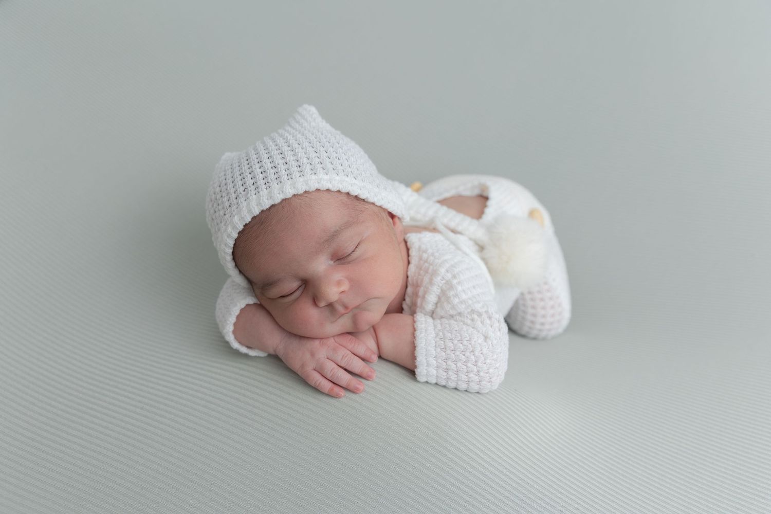 A sleeping newborn wearing a white knit outfit lies peacefully on a gray background.