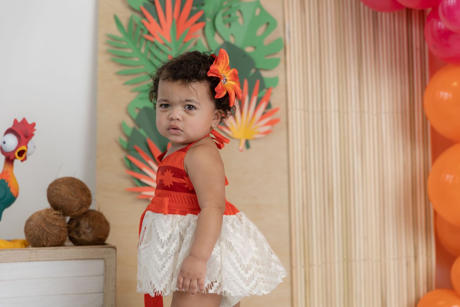 A toddler in tropical-themed dress poses in front of colorful party decorations.