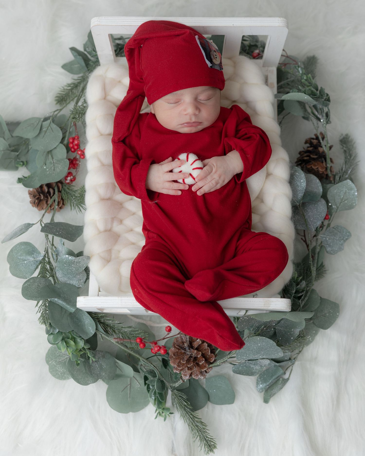 A sleeping baby in red holiday outfit rests on white bed surrounded by greenery and pinecones.