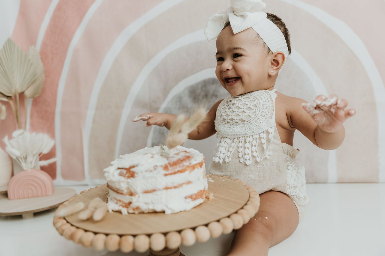 A baby in white lace outfit sits excitedly next to first birthday cake on decorative stand.