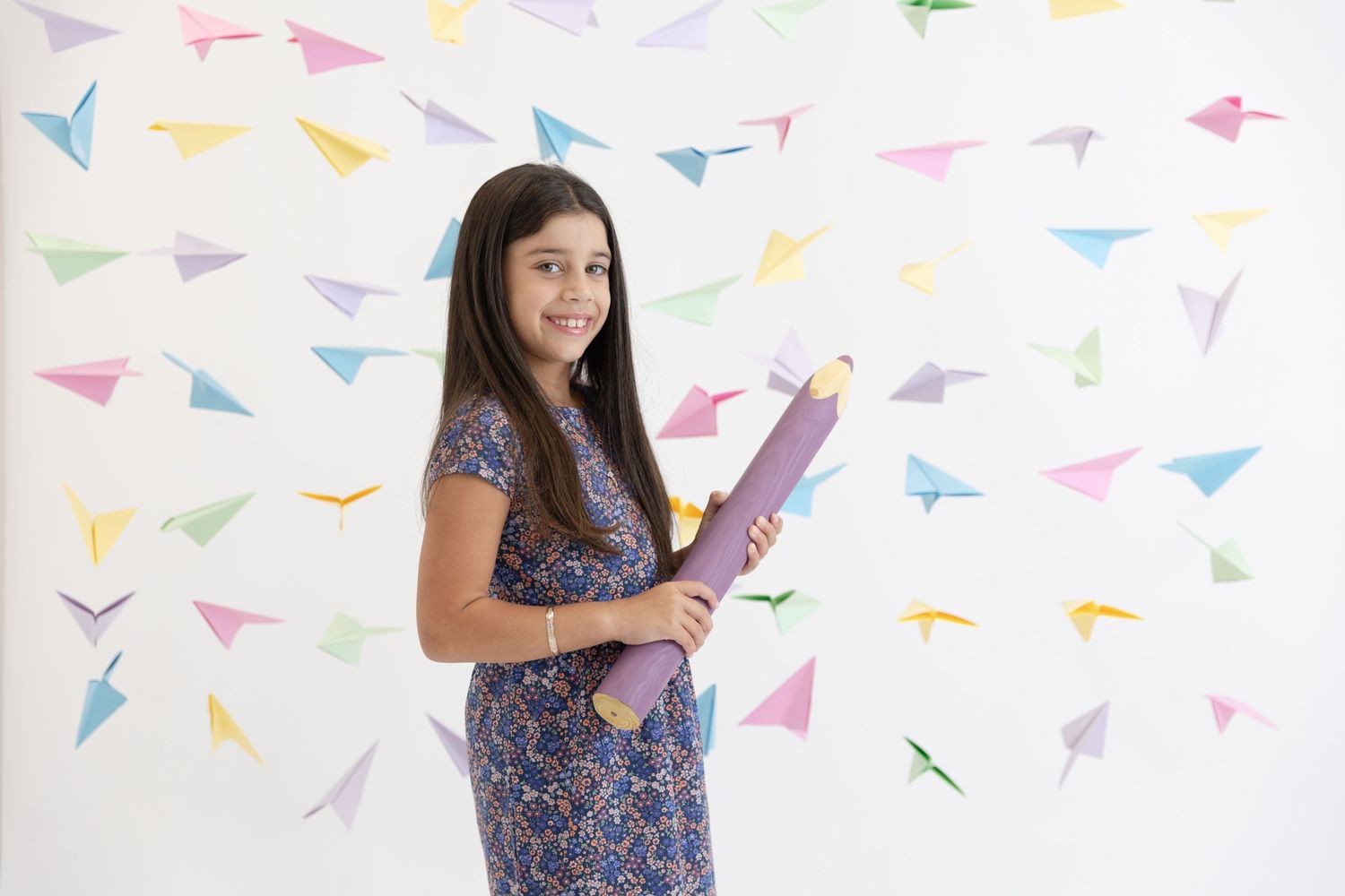 Student in floral dress holding large purple pencil prop against paper airplane pattern backdrop.