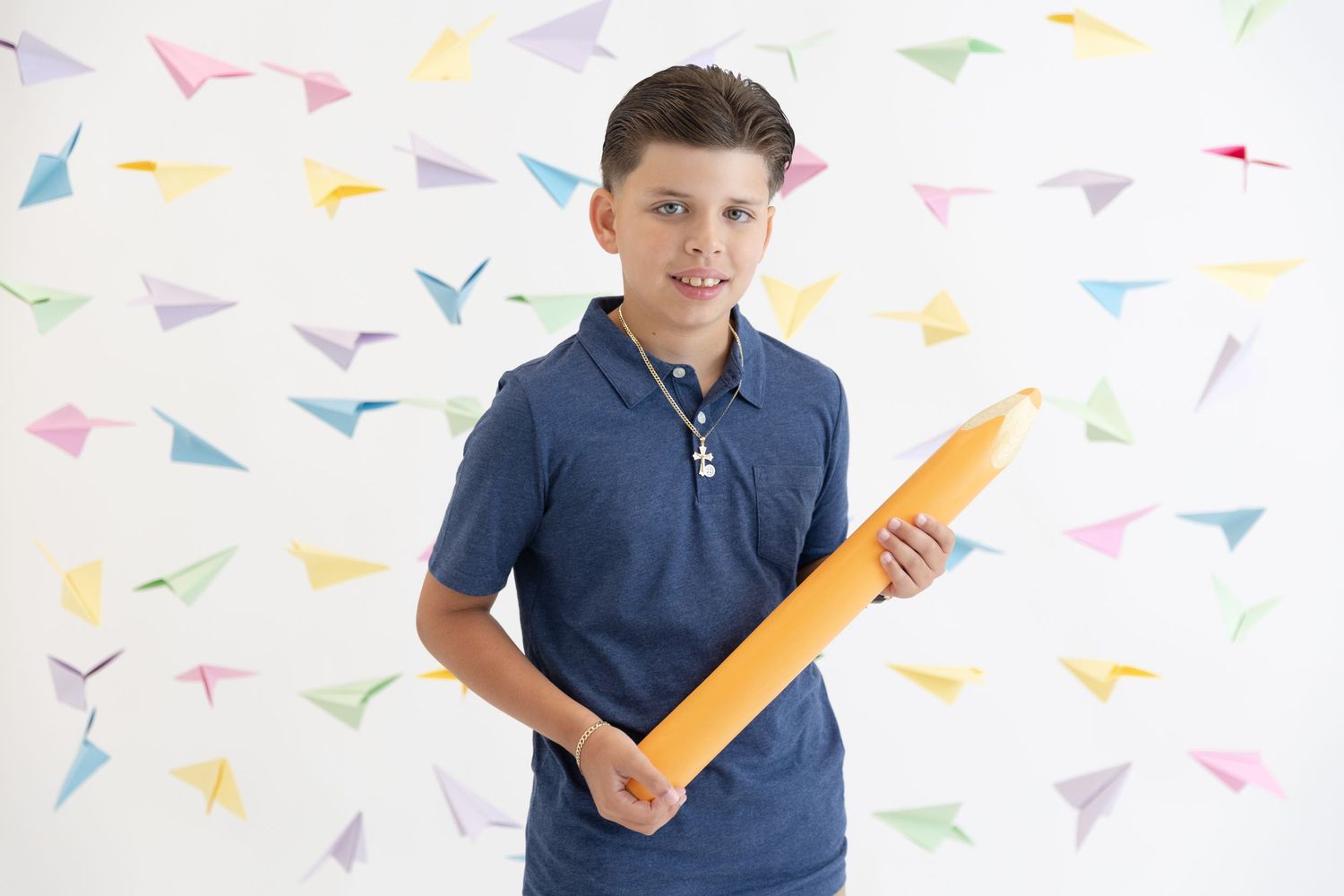 Young student holding large orange pencil prop against colorful paper airplane background.