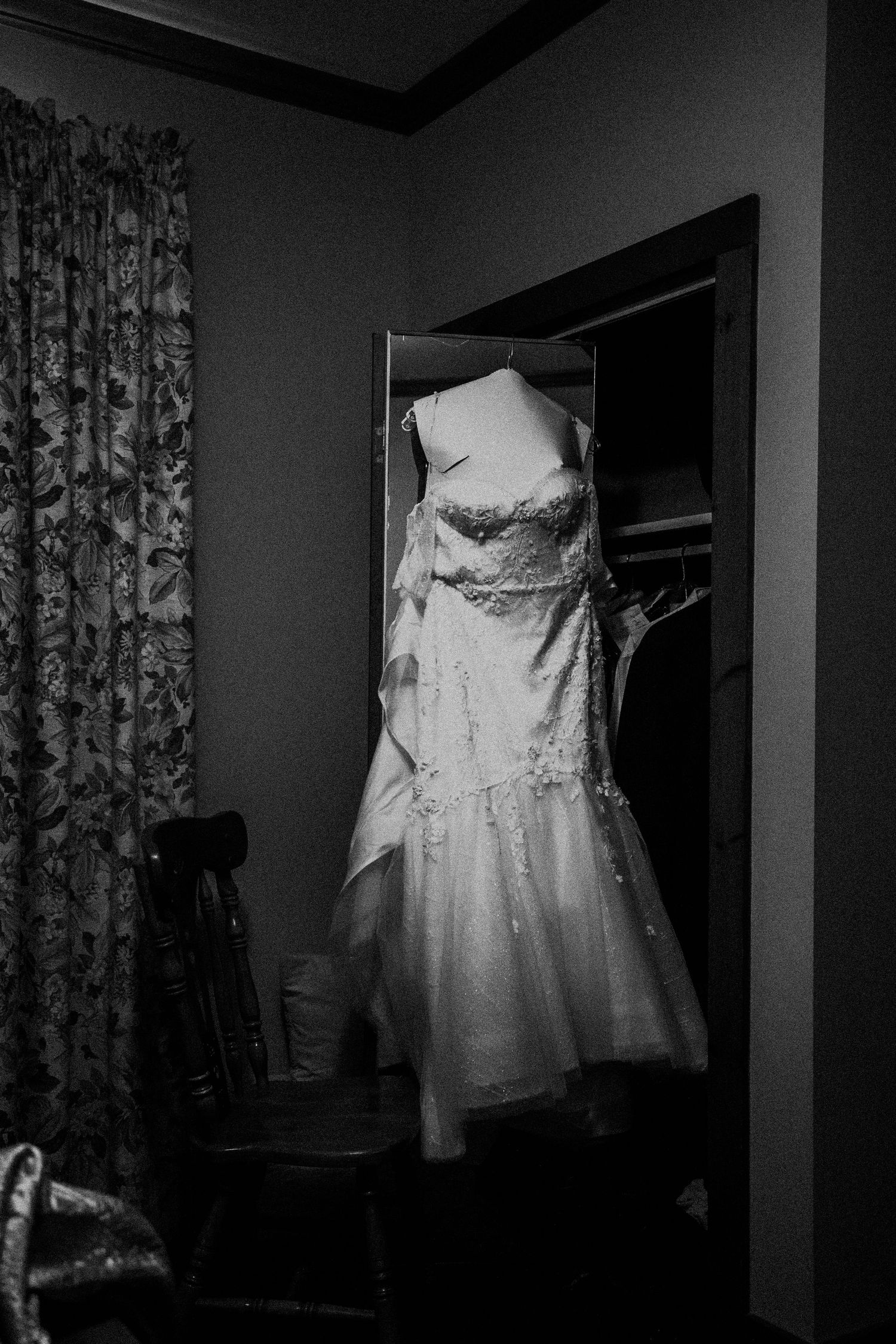 A white wedding dress hangs elegantly in a doorway against dark curtains in black and white.