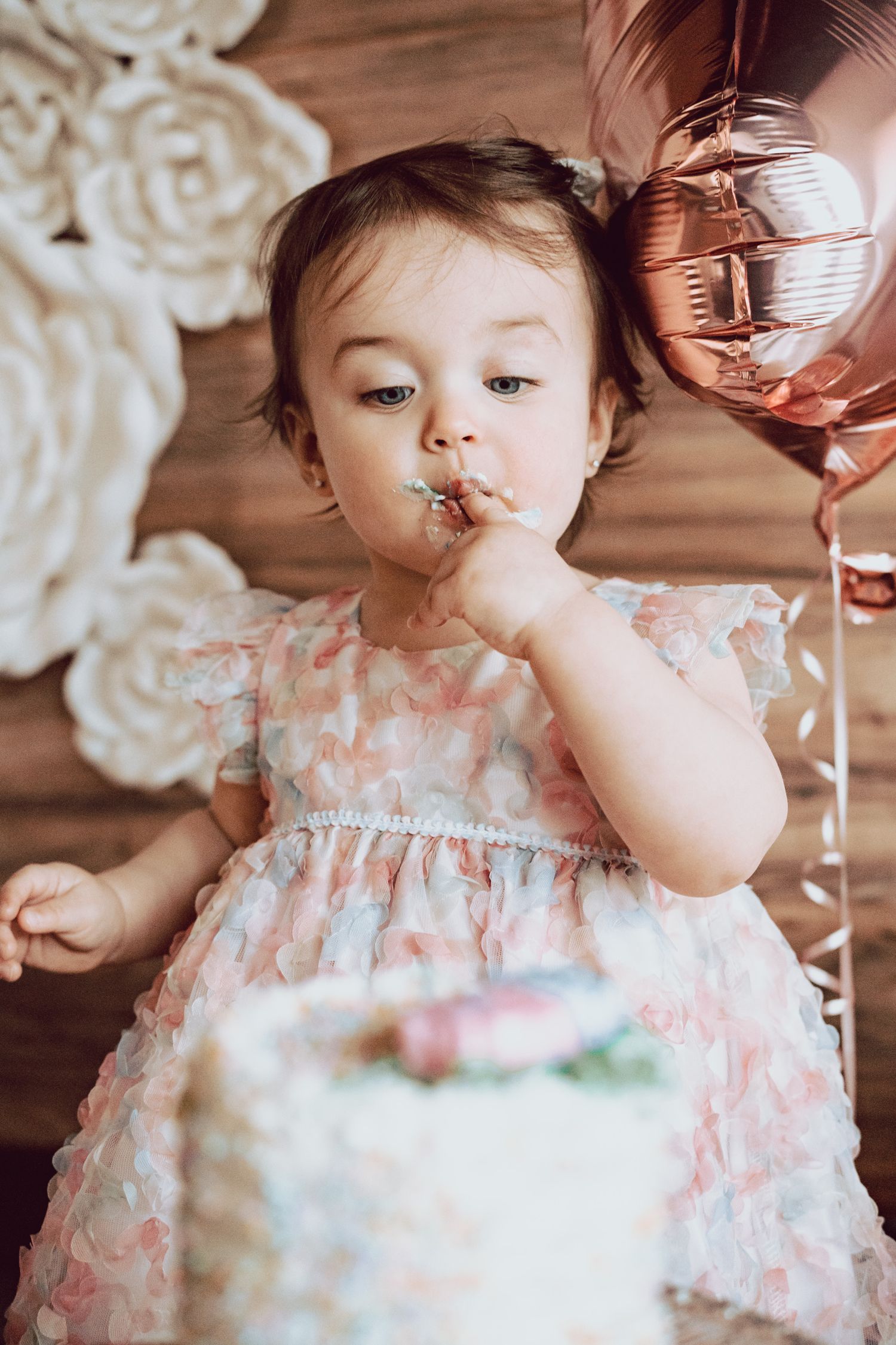 Toddler girl in floral dress eating birthday cake with rose gold balloons and paper flower backdrop.